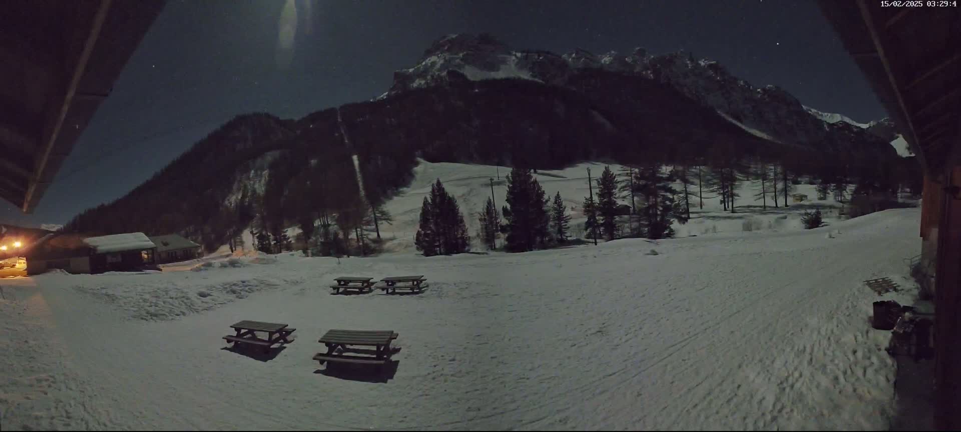 Under a moonlit night sky, several picnic tables sit in a snow-covered area in front of a snow-dusted mountain and some small buildings.