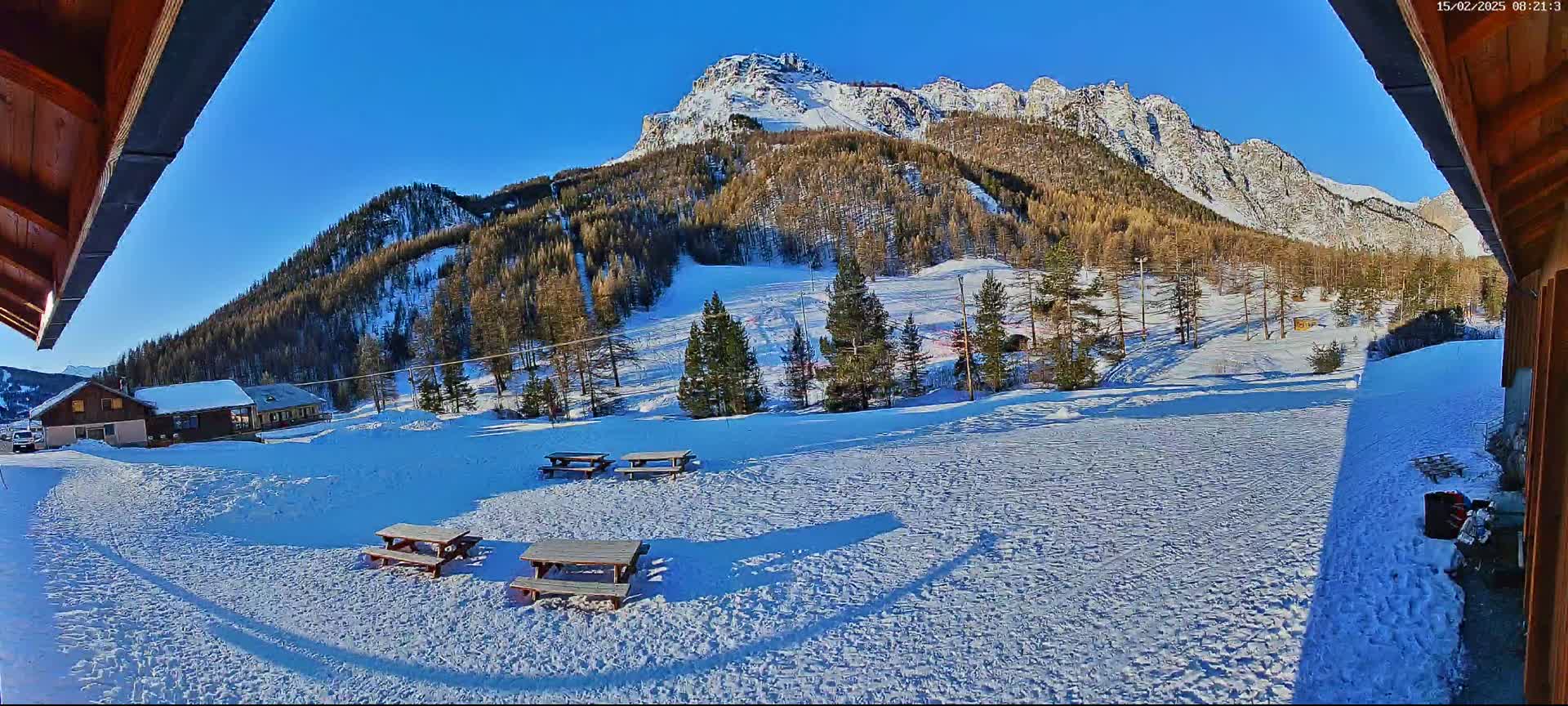 A snow-covered landscape with several picnic tables, a cluster of buildings, and a mountain range in the background under a clear blue sky.