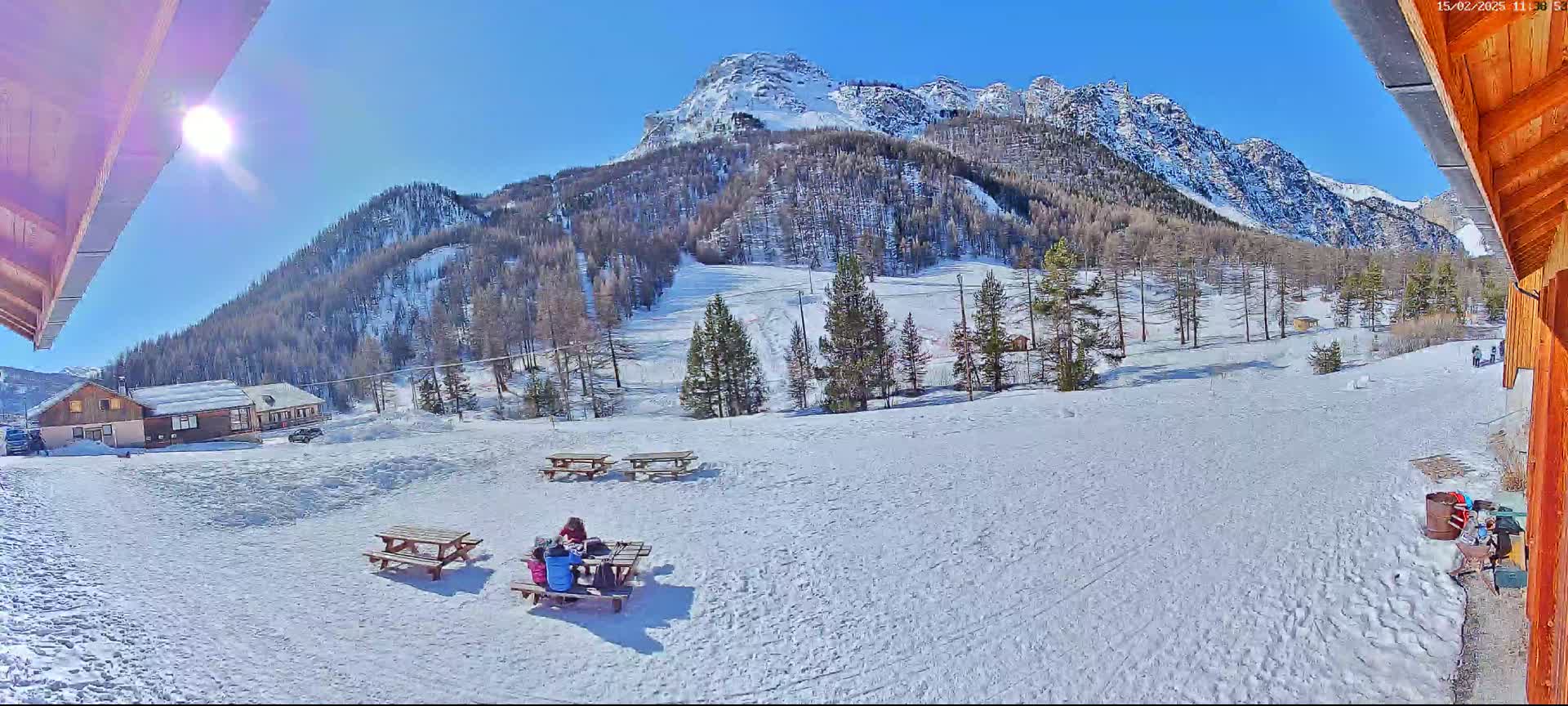 A sunny, snowy mountainside scene features several picnic tables, with a group of people seated at one, overlooking a snow-covered landscape and a distant mountain peak.
