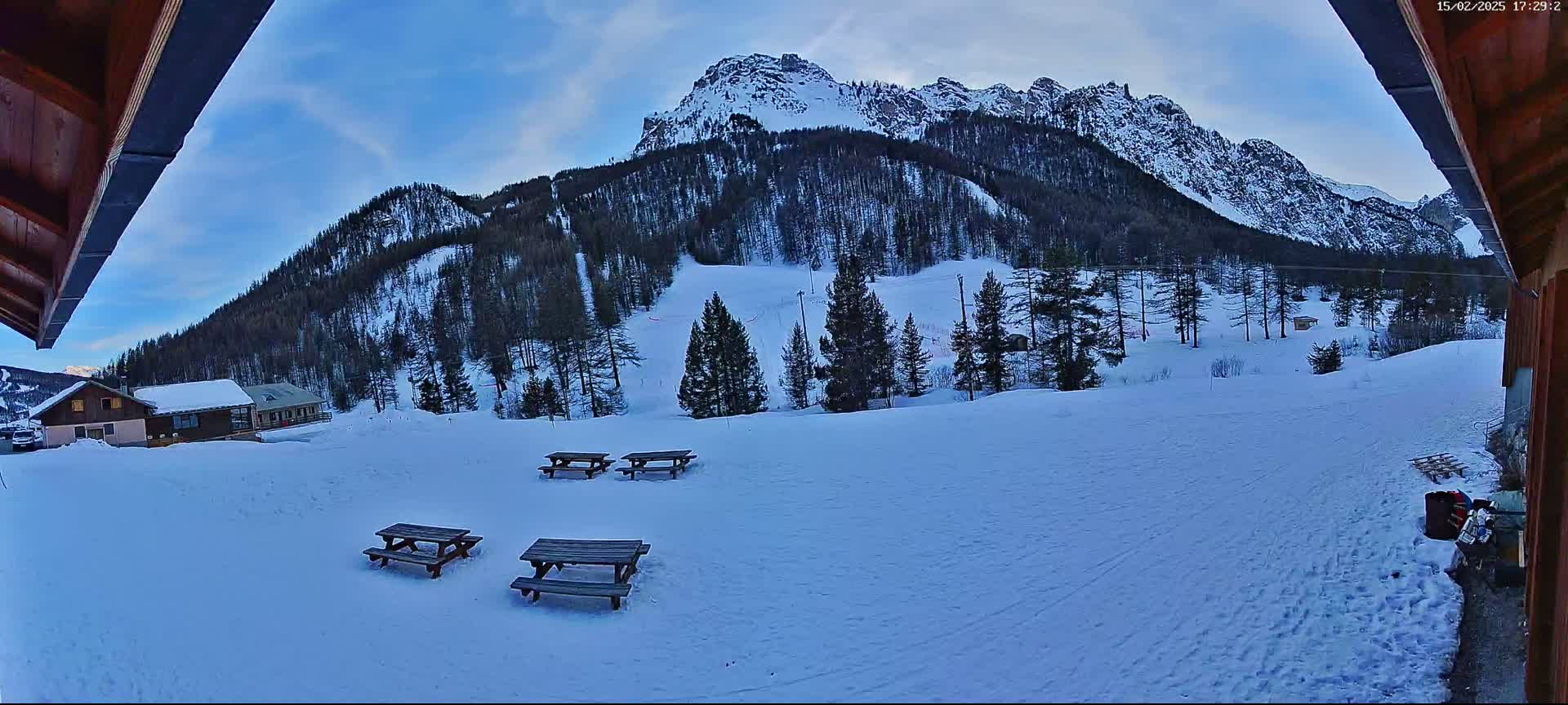 A snow-covered landscape with several picnic tables, a cluster of buildings, and a large mountain in the background under a partly cloudy sky.