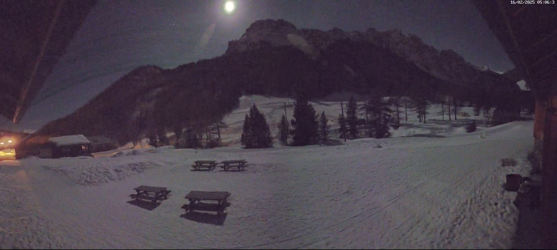 A moonlit snowy landscape at night features a few picnic tables in the foreground, some buildings, and a mountain in the background.