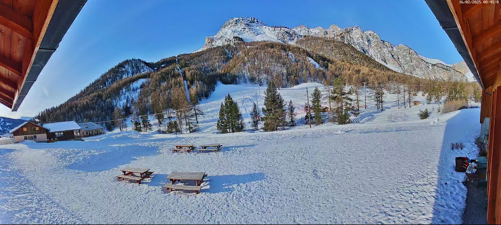 A snow-covered mountainside with several picnic tables and small buildings is visible under a clear blue sky.