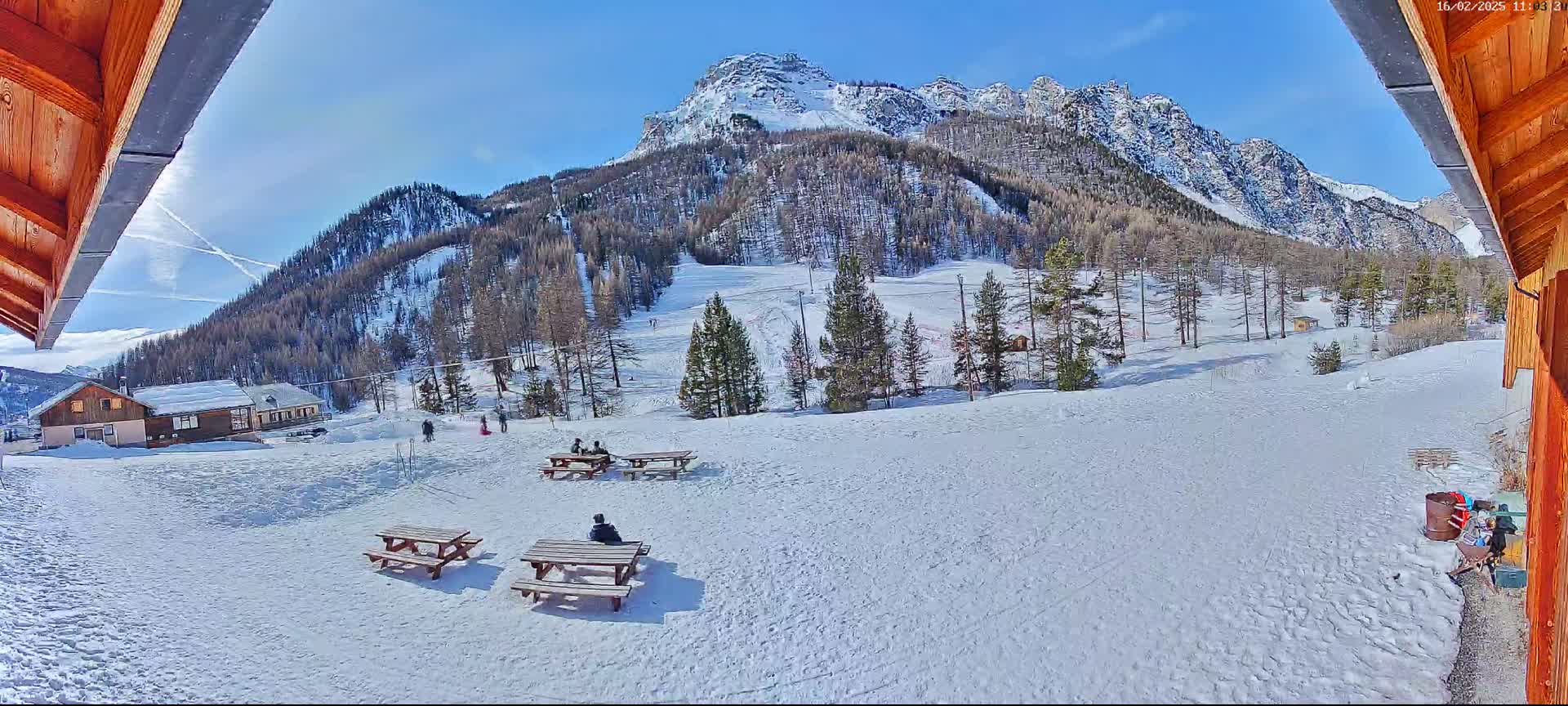 A snowy landscape with picnic tables, a few people, buildings, and a mountain range in the background under a sunny sky.