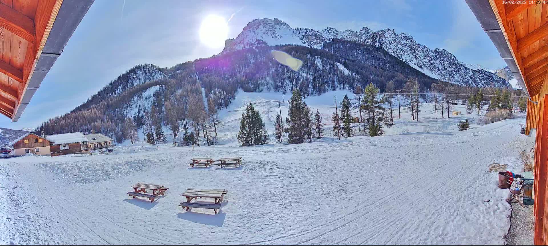 A sunny winter scene shows snow-covered picnic tables and a ski slope below a mountain range viewed from a wooden structure.