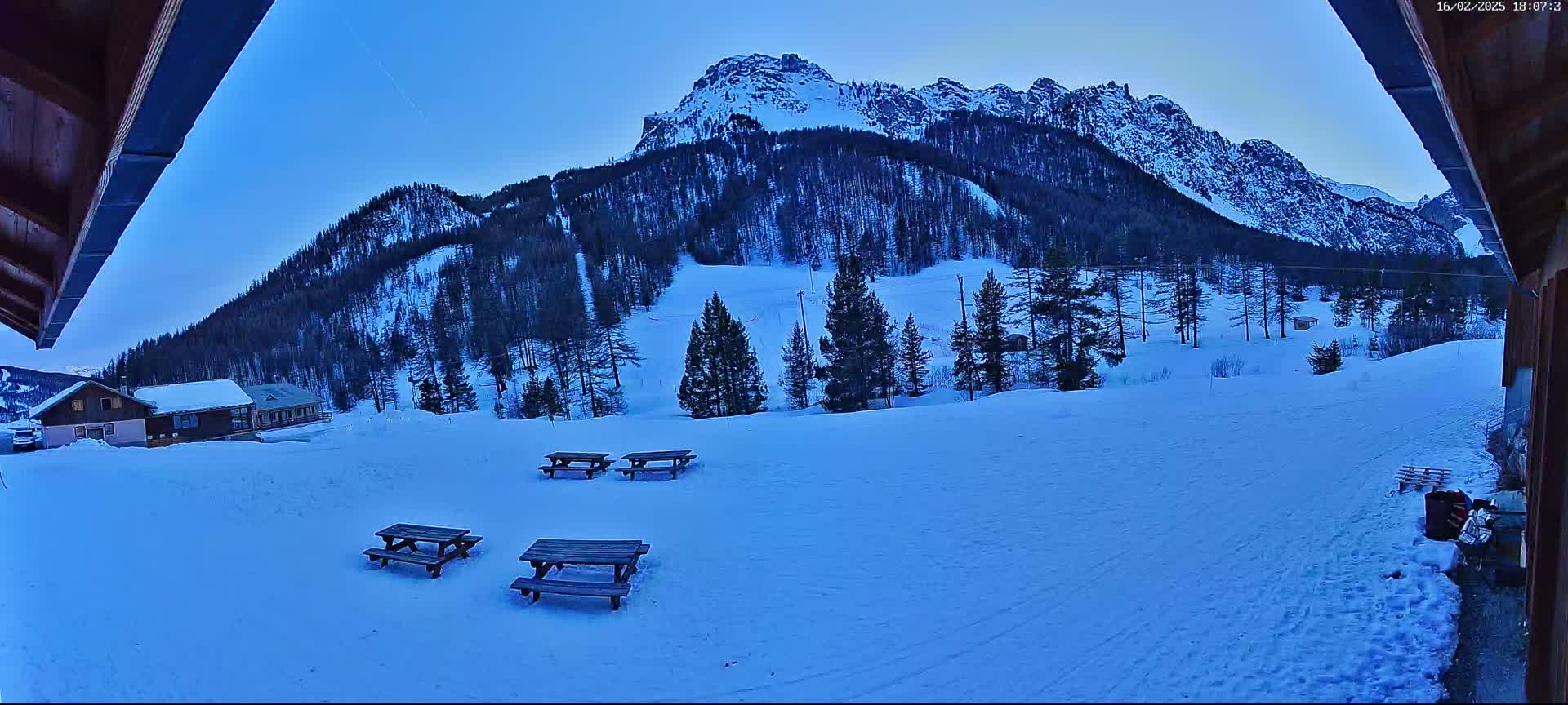 A snow-covered landscape with several picnic tables, a few small buildings, and a large mountain in the background under a twilight sky.