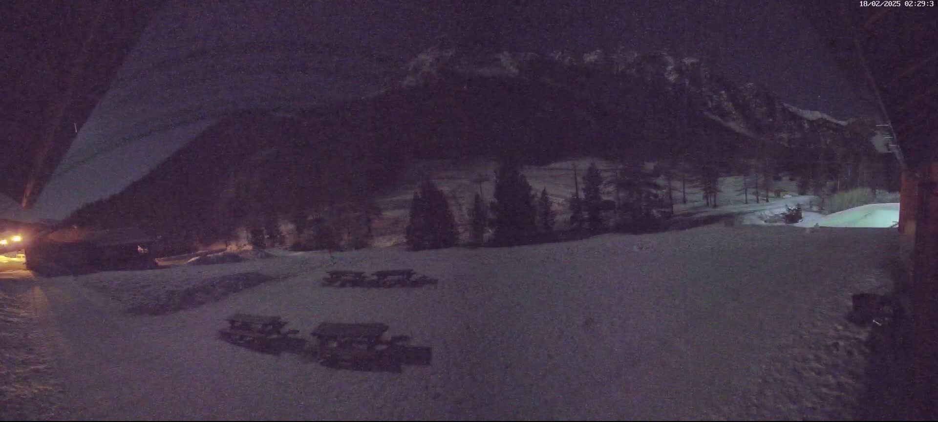 A nighttime view of a snow-covered area with picnic tables, trees, and mountains in the background under a dark sky.
