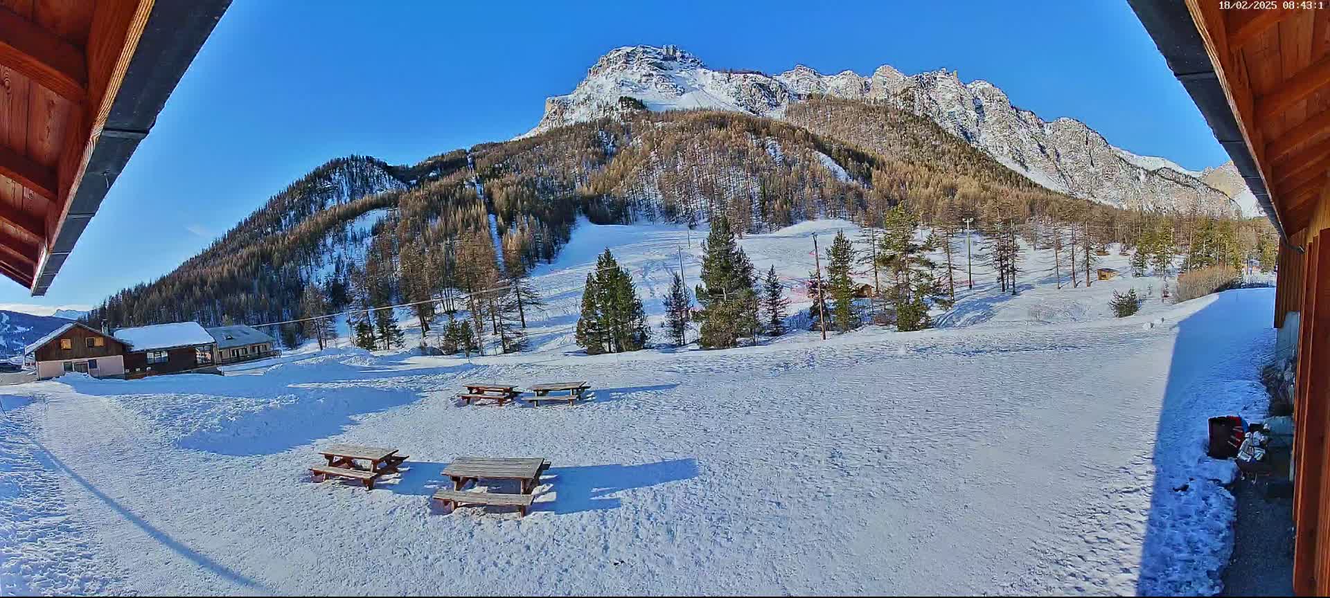 A snow-covered landscape with picnic tables sits in front of a mountain range under a clear blue sky.