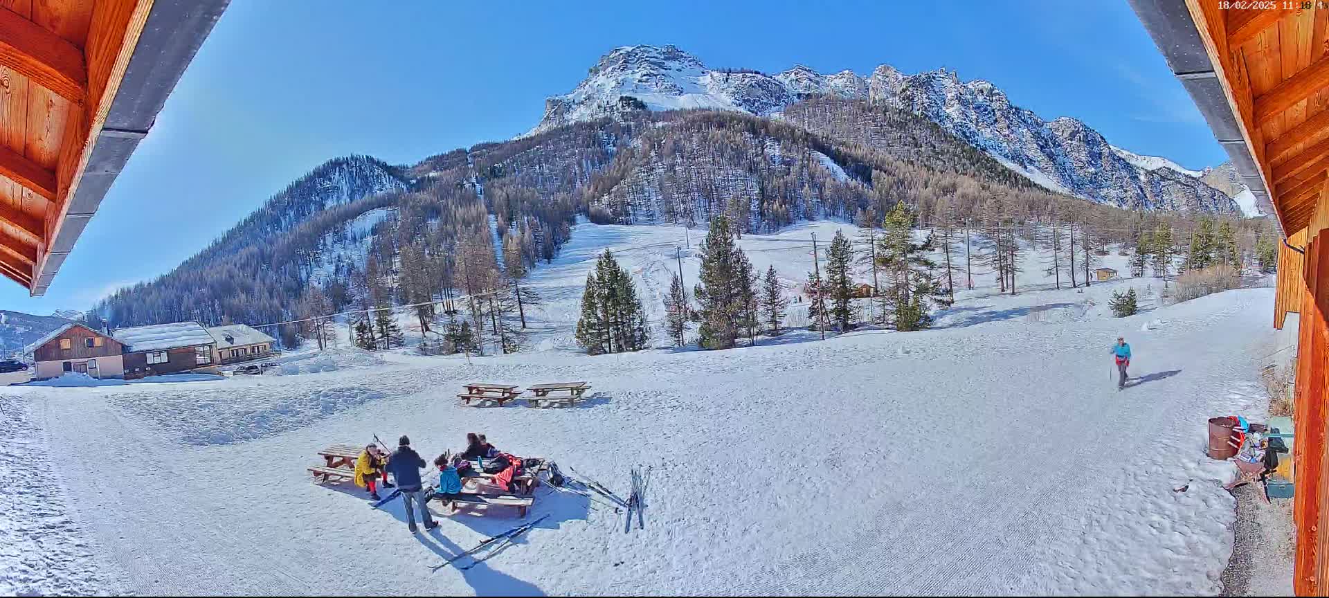 A sunny, snow-covered mountain landscape is viewed from a wooden structure, showing several people sitting at picnic tables and a lone person walking in the distance.