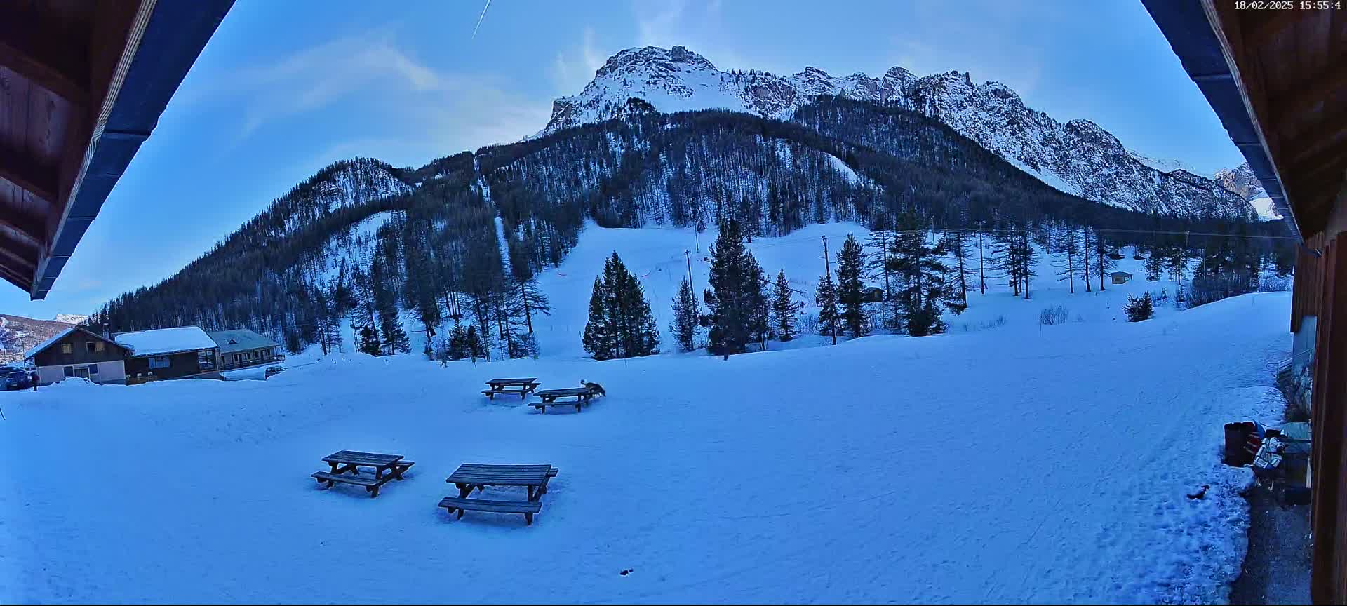 A snow-covered landscape with several picnic tables in the foreground and a snow-capped mountain in the background under a clear, blue sky.