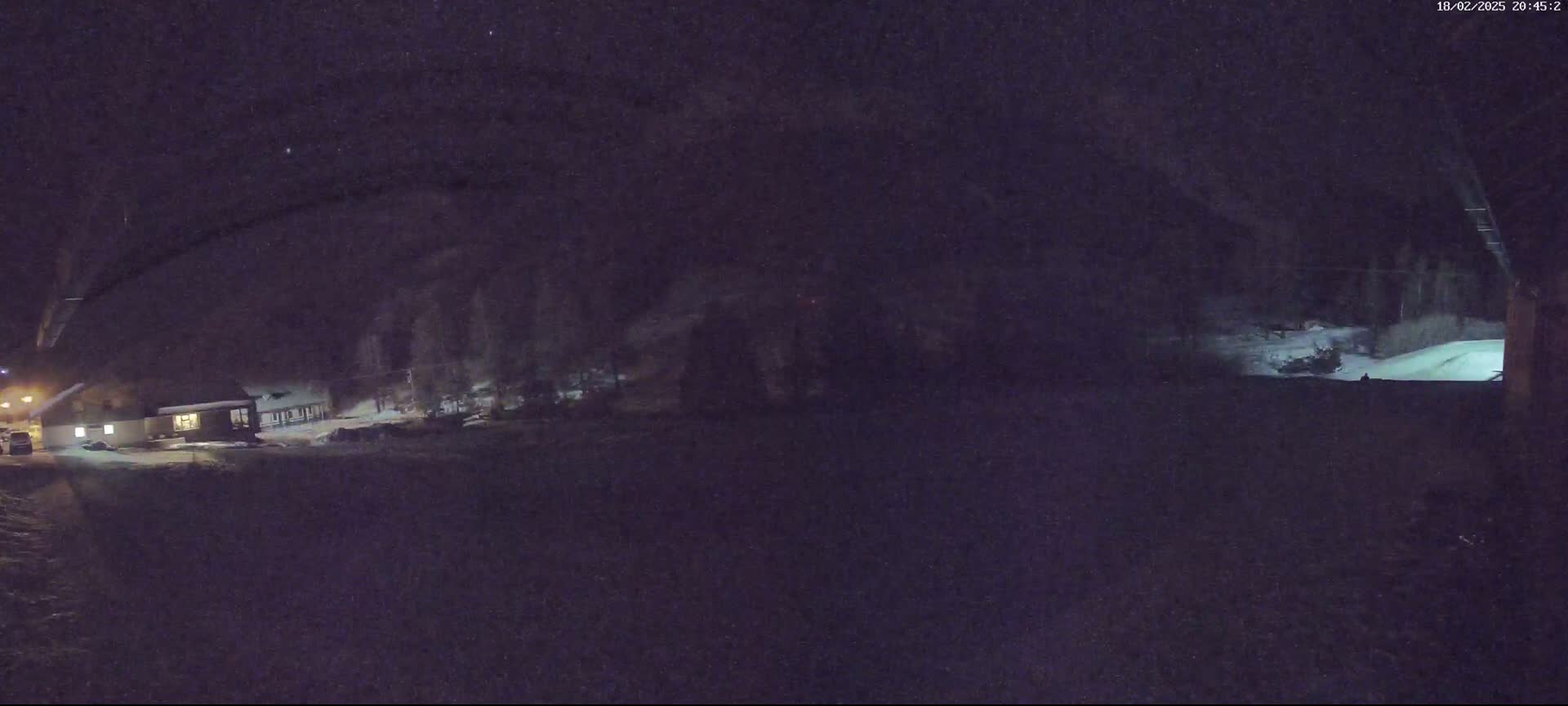 A nighttime, snow-covered scene shows a building with lights on, trees, and a partially visible flat area, all under a dark, starry sky.
