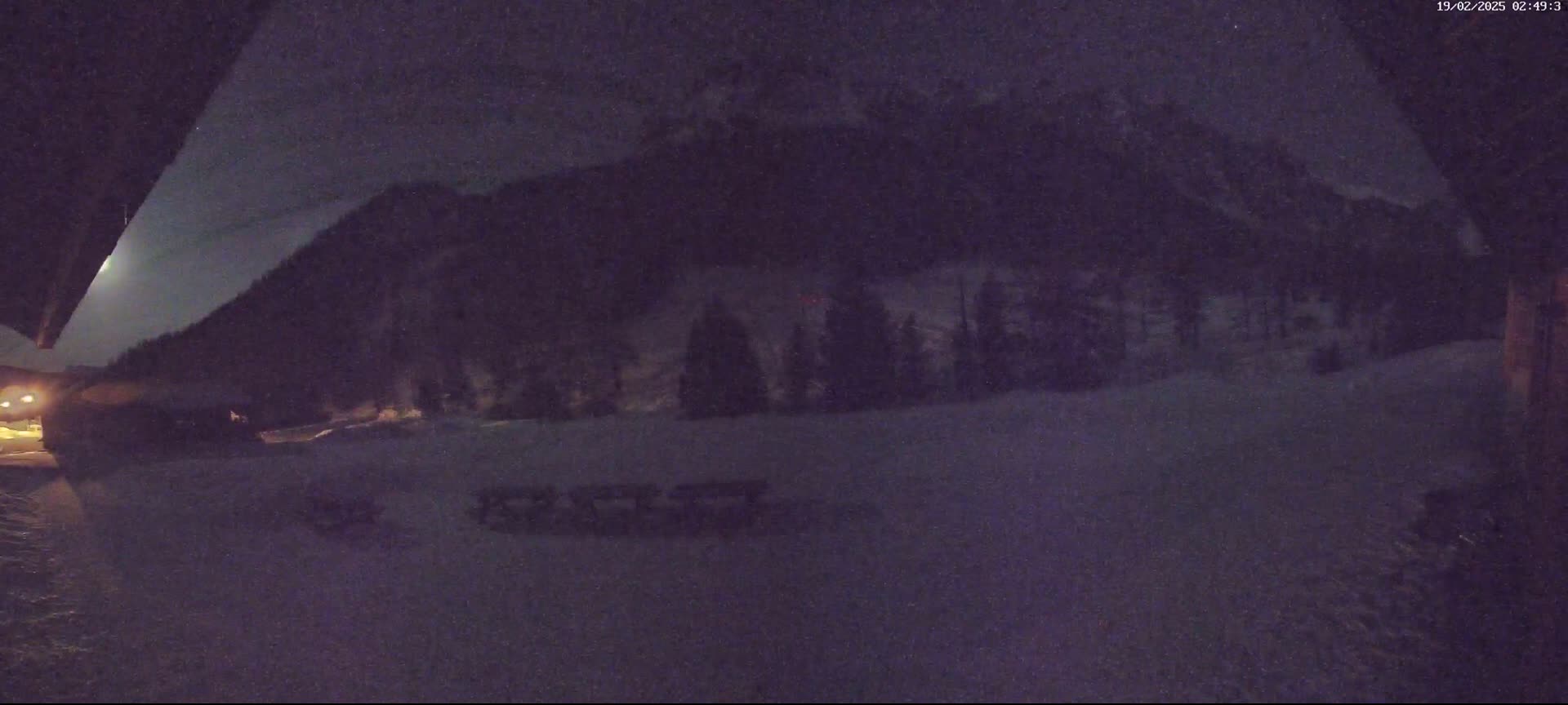 A nighttime, snow-covered landscape is seen from a building's edge, showing a moonlit mountain range and a line of dark trees in the middle ground.