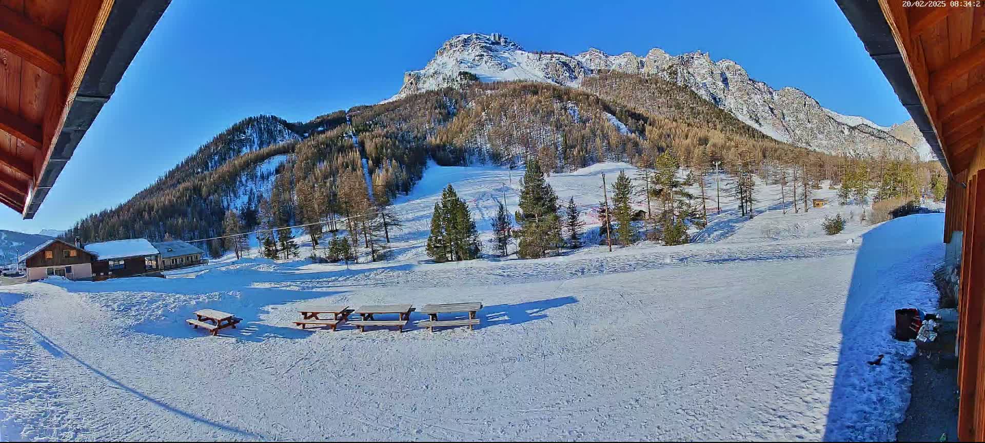 A snow-covered landscape with several picnic tables sits in the foreground, overlooking a snow-covered mountainside with sparsely populated evergreen trees under a clear blue sky.