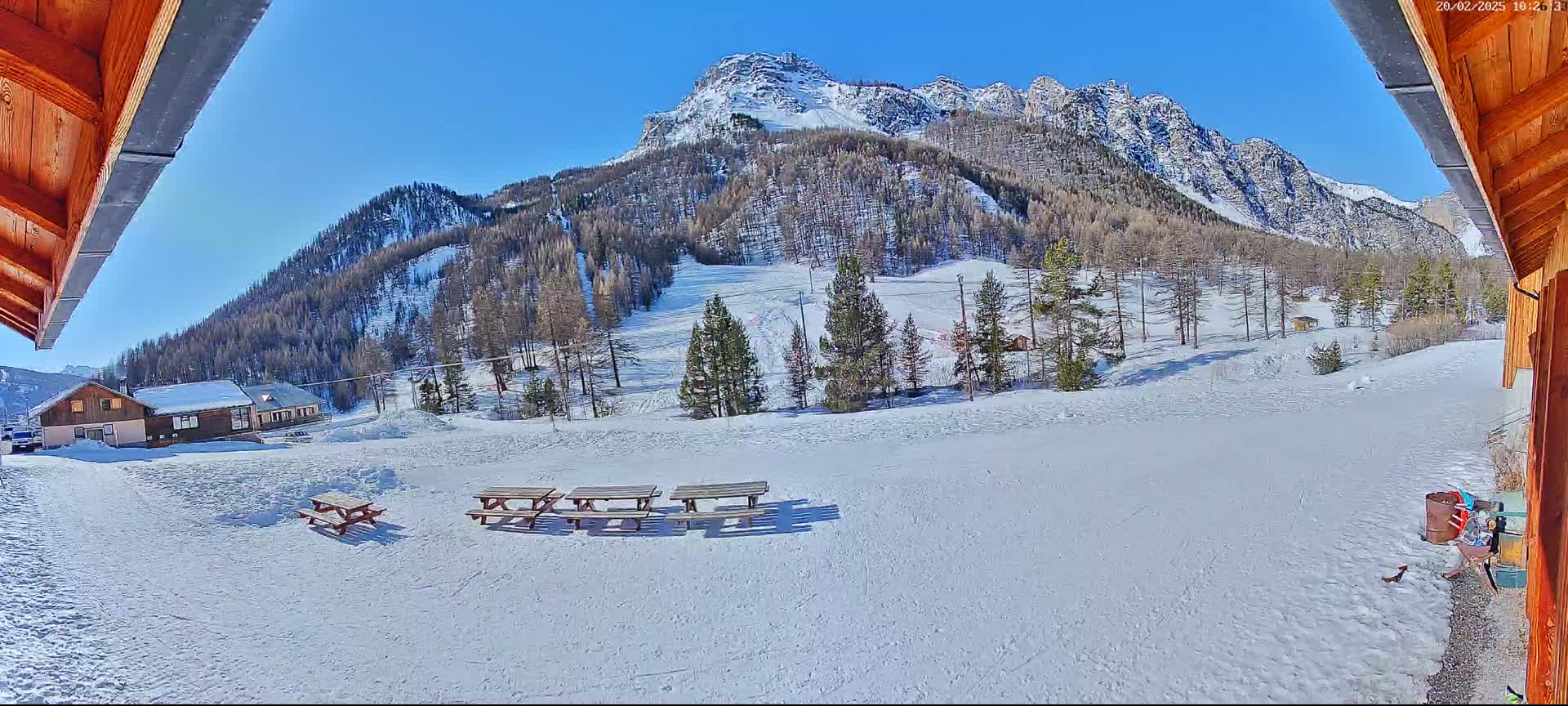 A sunny, snow-covered landscape features a mountainous backdrop, several picnic tables in the foreground, and a few buildings nestled among the trees.