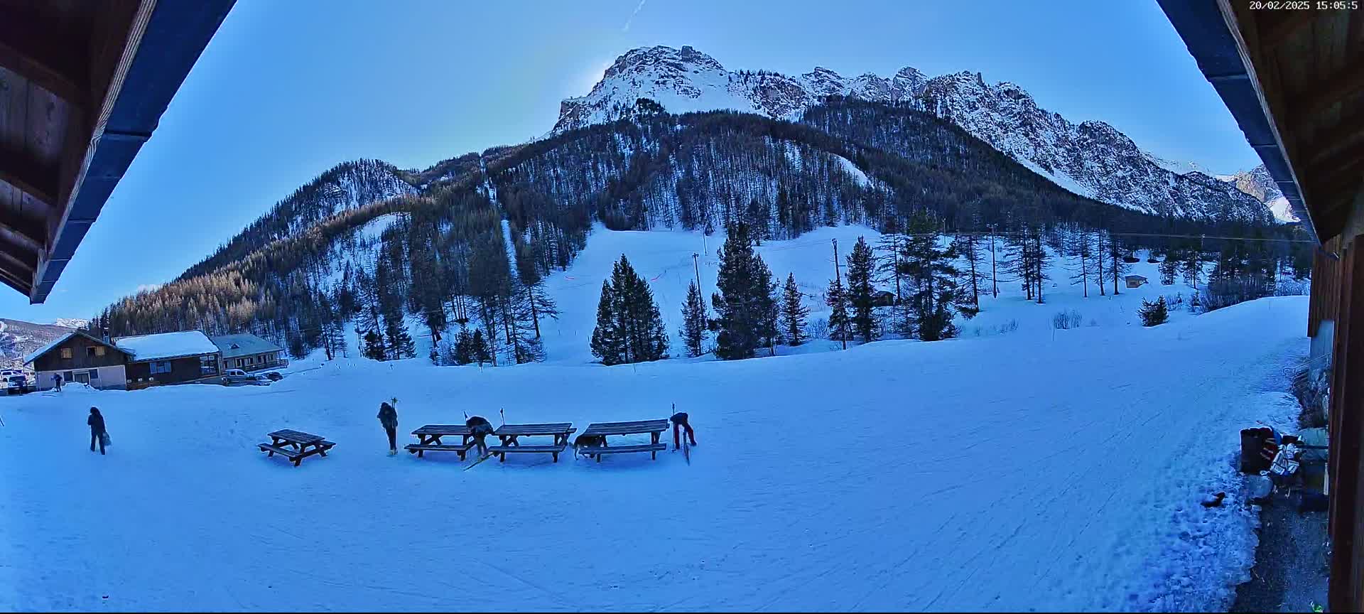 A snow-covered mountainside with several people near picnic tables, viewed from a partially covered outdoor area on a sunny day.