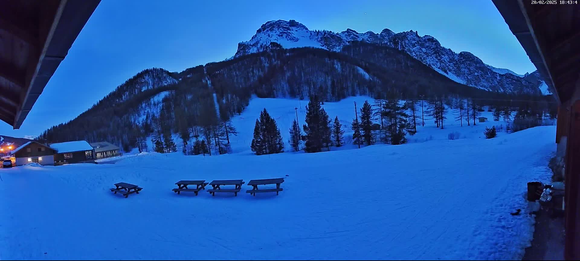 A snow-covered landscape at twilight, featuring a mountain range in the background, a few small buildings, and several picnic tables in the foreground.