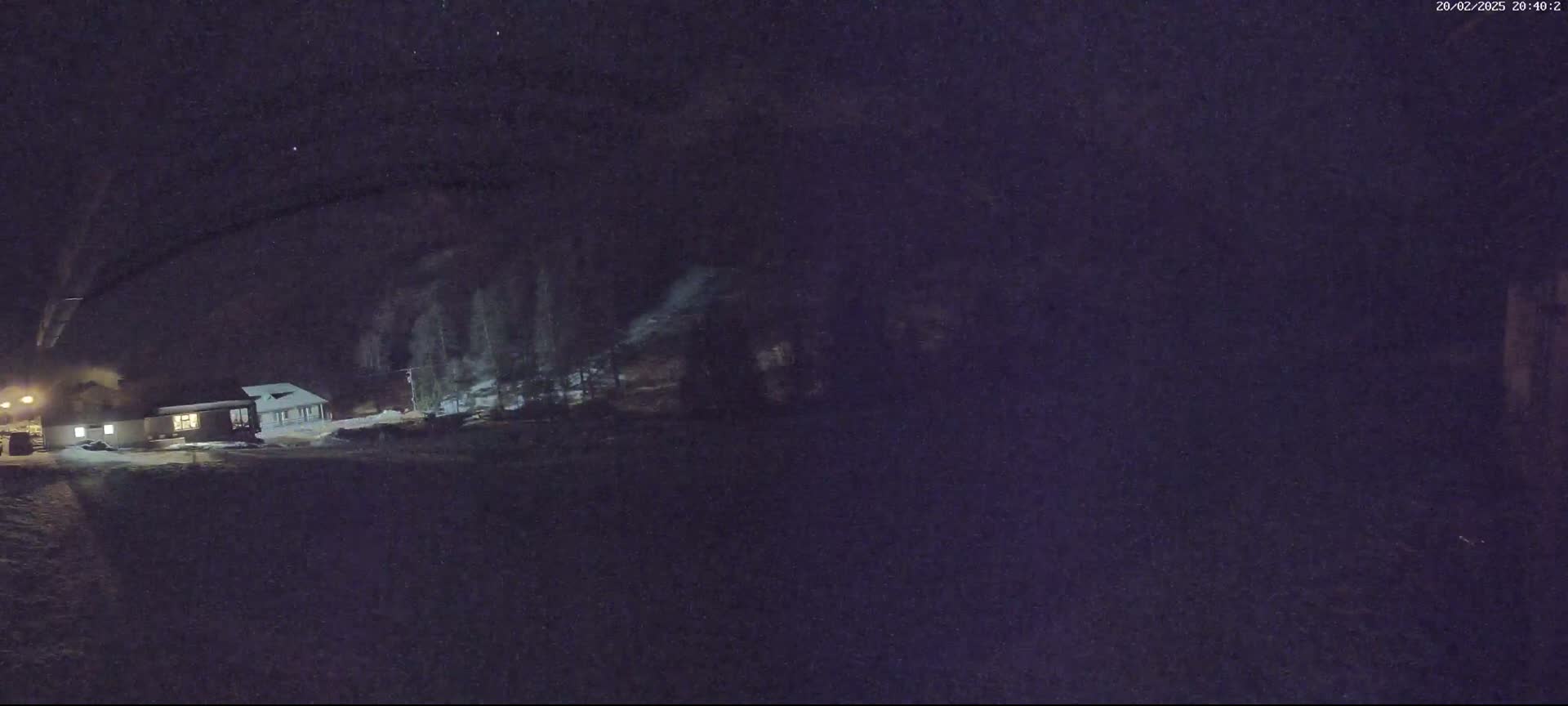 A low-light, nighttime image shows a building and some trees under a dark, clear sky with snow on the ground.