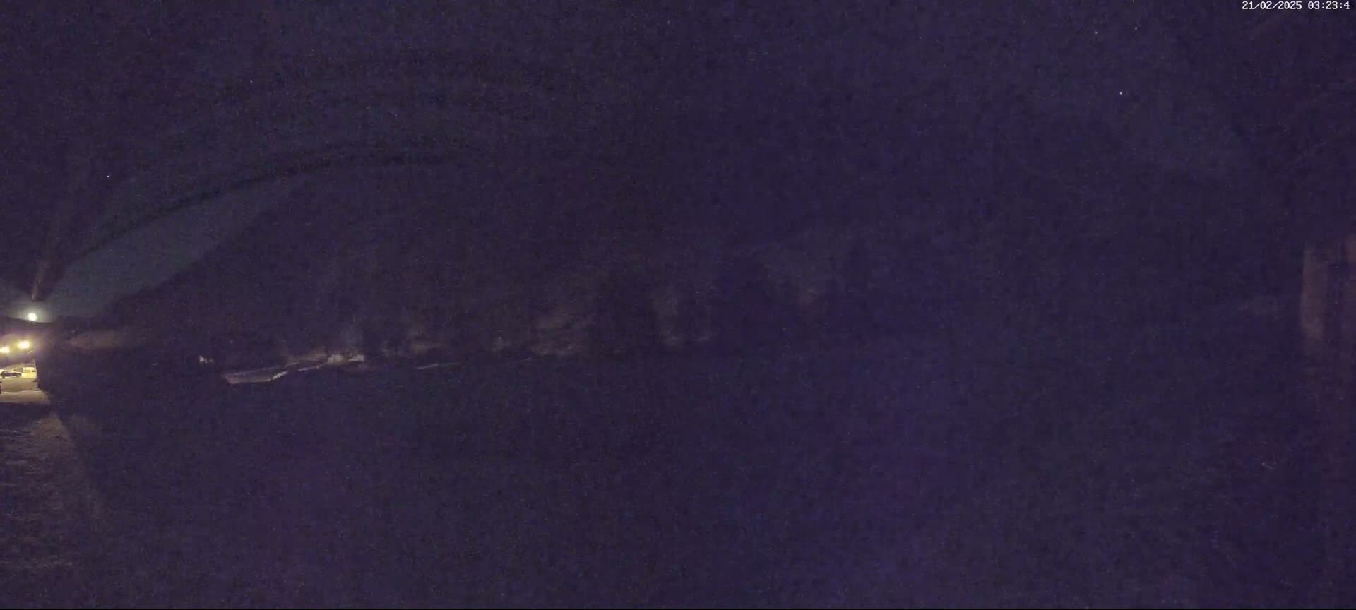 A dark, nighttime scene shows a dimly lit street with a few vehicles and some indistinct objects in the background under a clear, dark sky.
