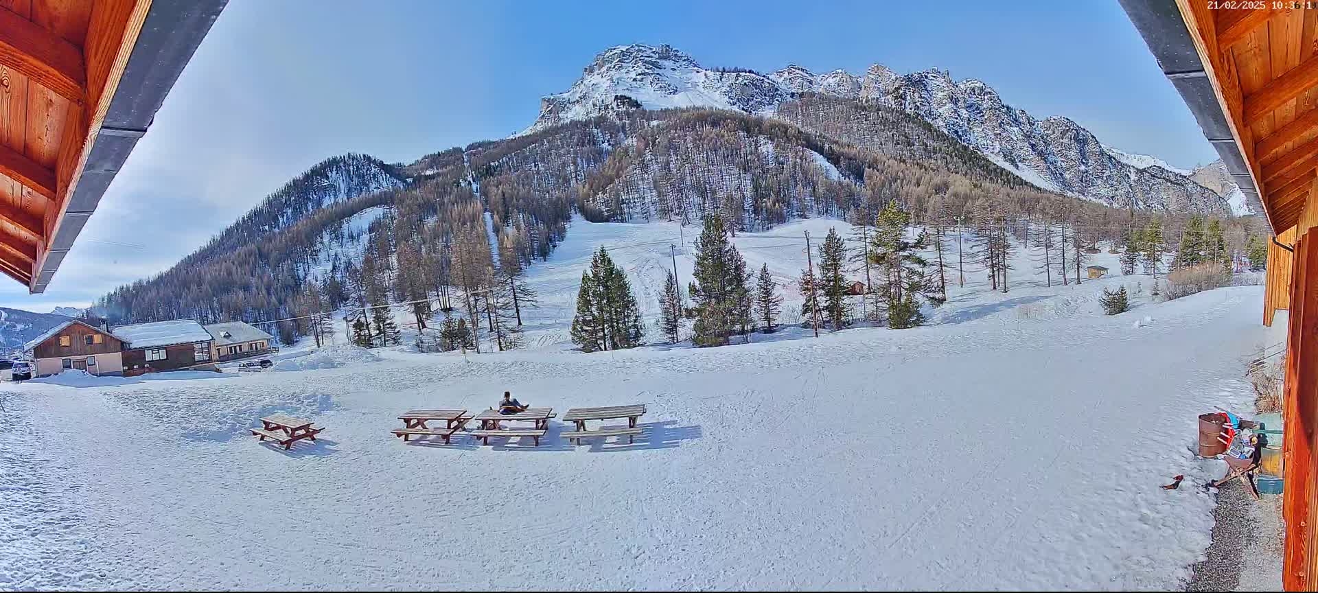 A snowy mountainside with several picnic tables, a person sitting at one, and a small building in the distance under a sunny sky.