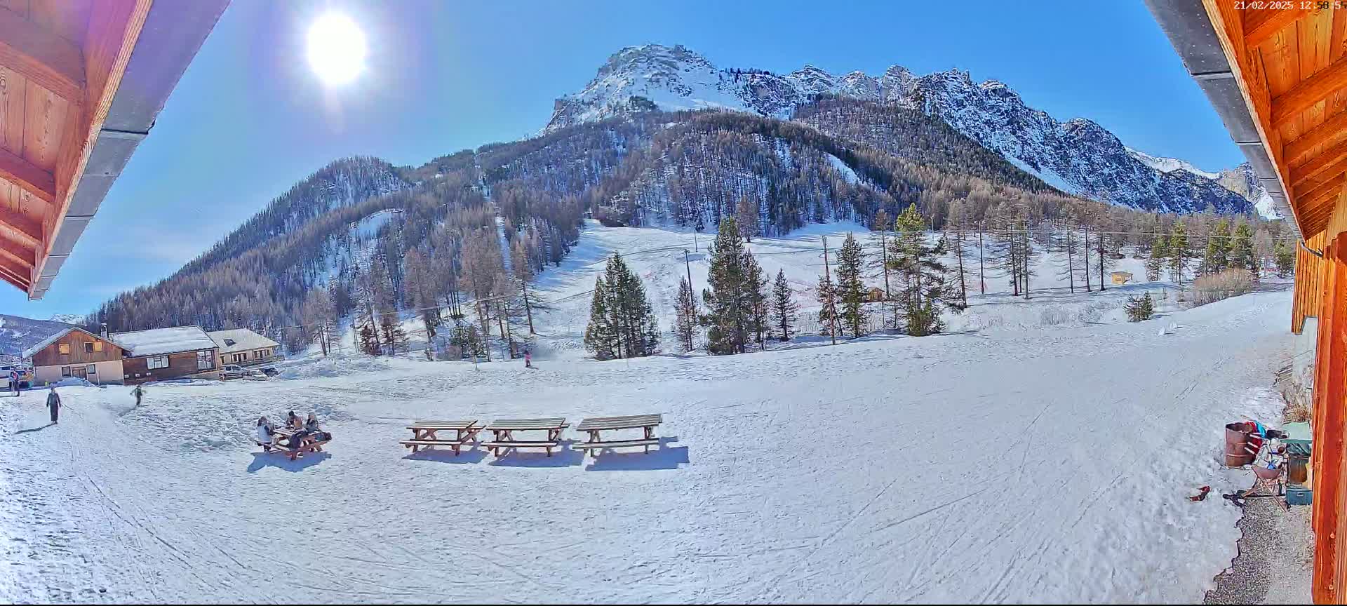 A sunny winter scene shows snow-covered ground with picnic tables, a few people, and buildings, all set against a backdrop of snow-capped mountains and evergreen trees.