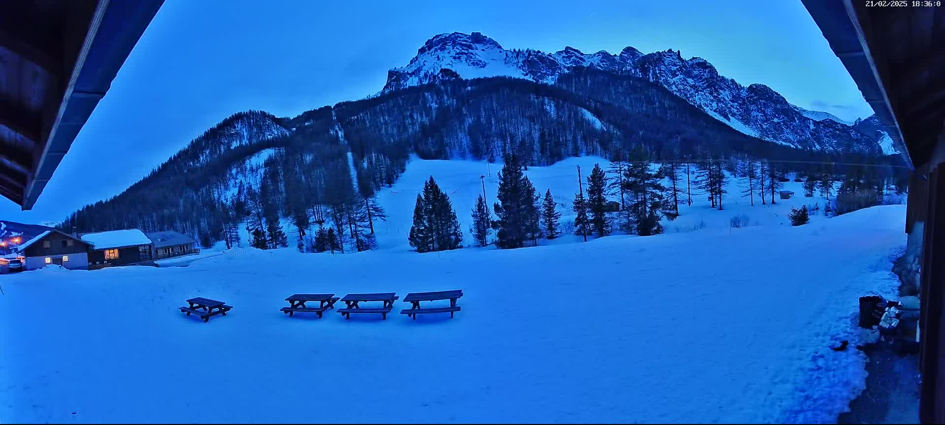 A snow-covered landscape at twilight, featuring a mountain range in the background, several picnic tables in the foreground, and a small building nestled amongst evergreen trees.