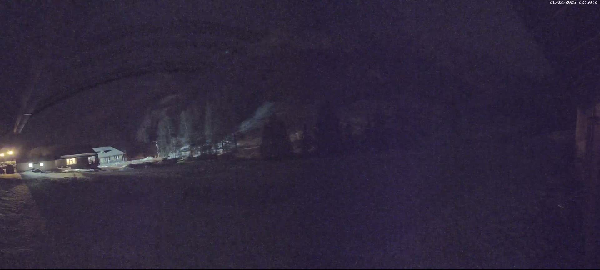 A low-light nighttime image shows a small house and some trees covered in snow.