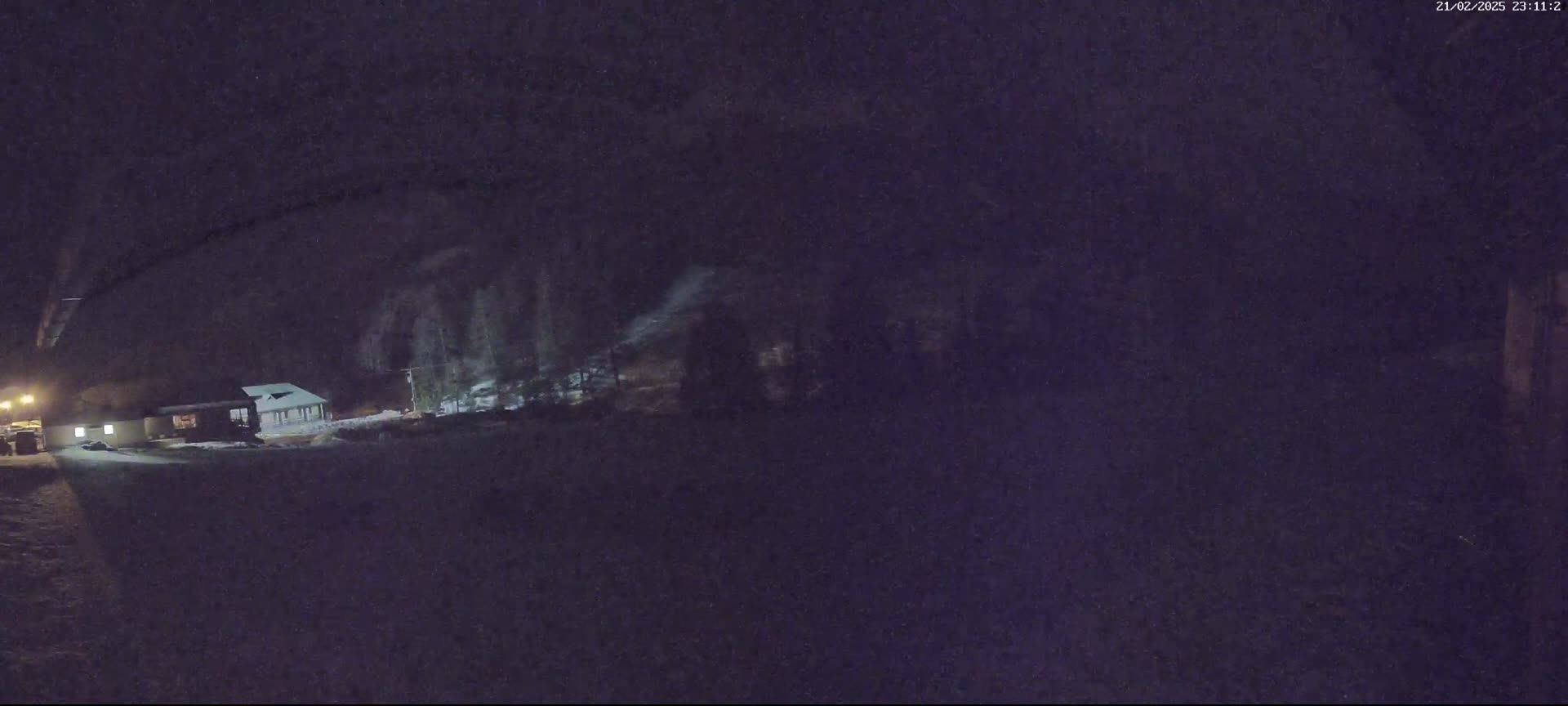 A dimly lit, snow-covered area at night shows a building partially obscured by trees and darkness.