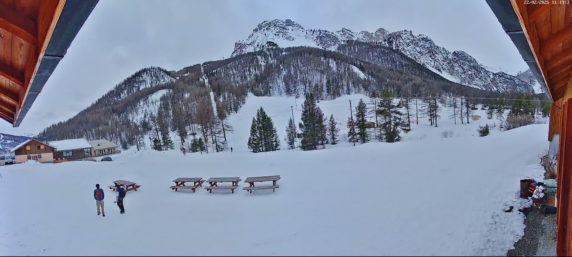 A snowy landscape with a mountain in the background, several picnic tables in the foreground, and two people walking toward a building.