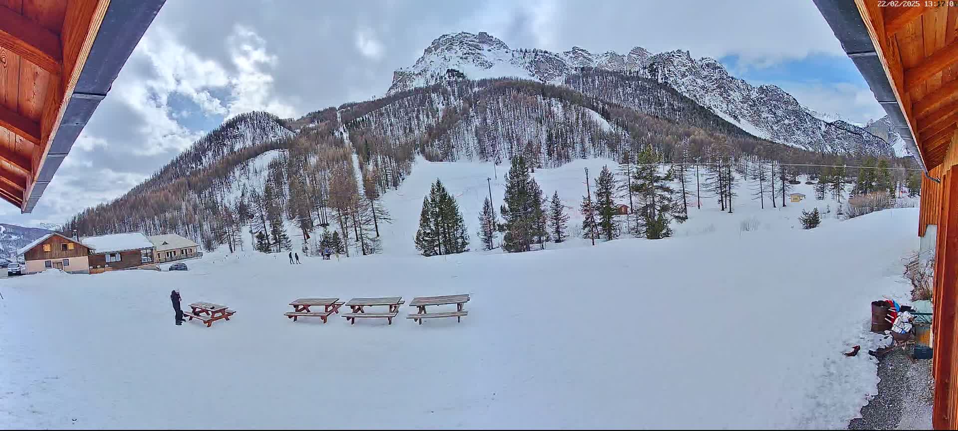 A snowy, mountainous landscape is viewed from under a wooden overhang, showing several picnic tables in the foreground and a few people in the distance.