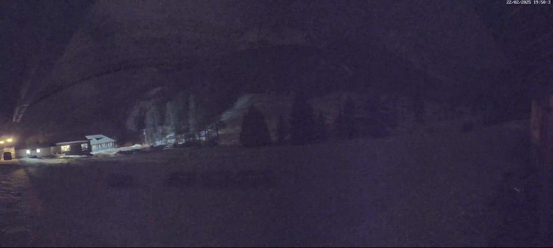 A snow-covered landscape at night shows a small building illuminated by artificial light, with dark trees behind it under a dark sky.