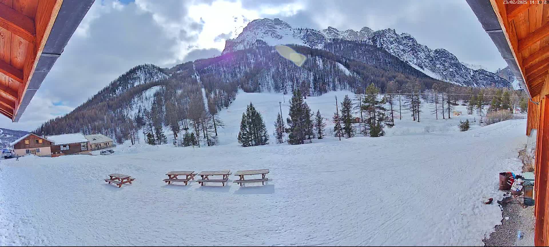 A snowy mountainside with several picnic tables in the foreground, viewed from under a wooden structure on a partly cloudy day.