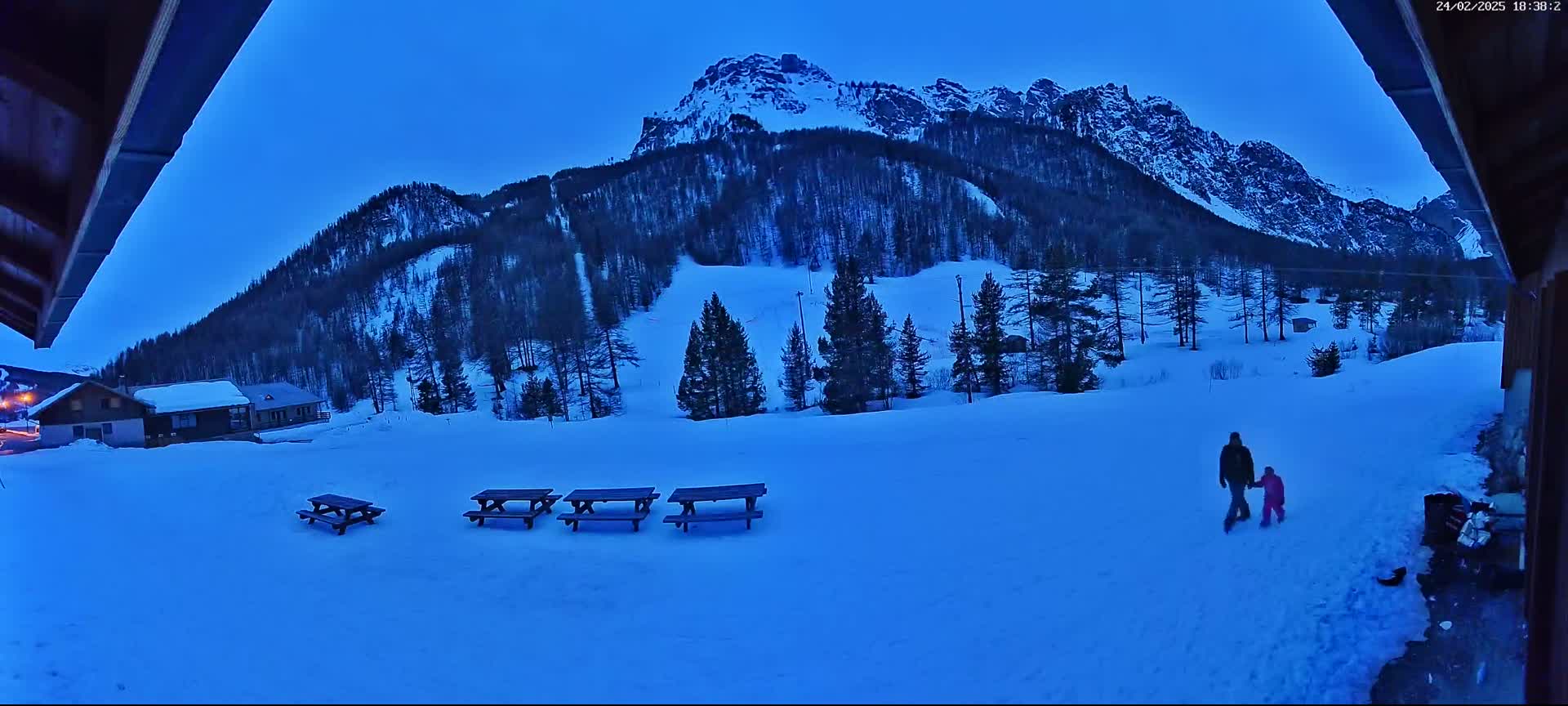 A snow-covered landscape at twilight, featuring a mountain range in the background, several picnic tables in the foreground, and two figures walking hand-in-hand.