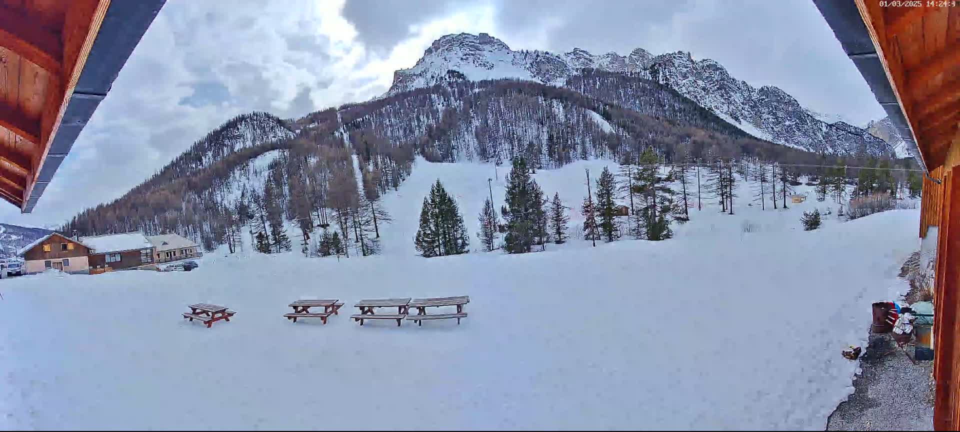 A snow-covered landscape featuring a mountain range under a partly cloudy sky, several picnic tables, and a small building in the foreground.