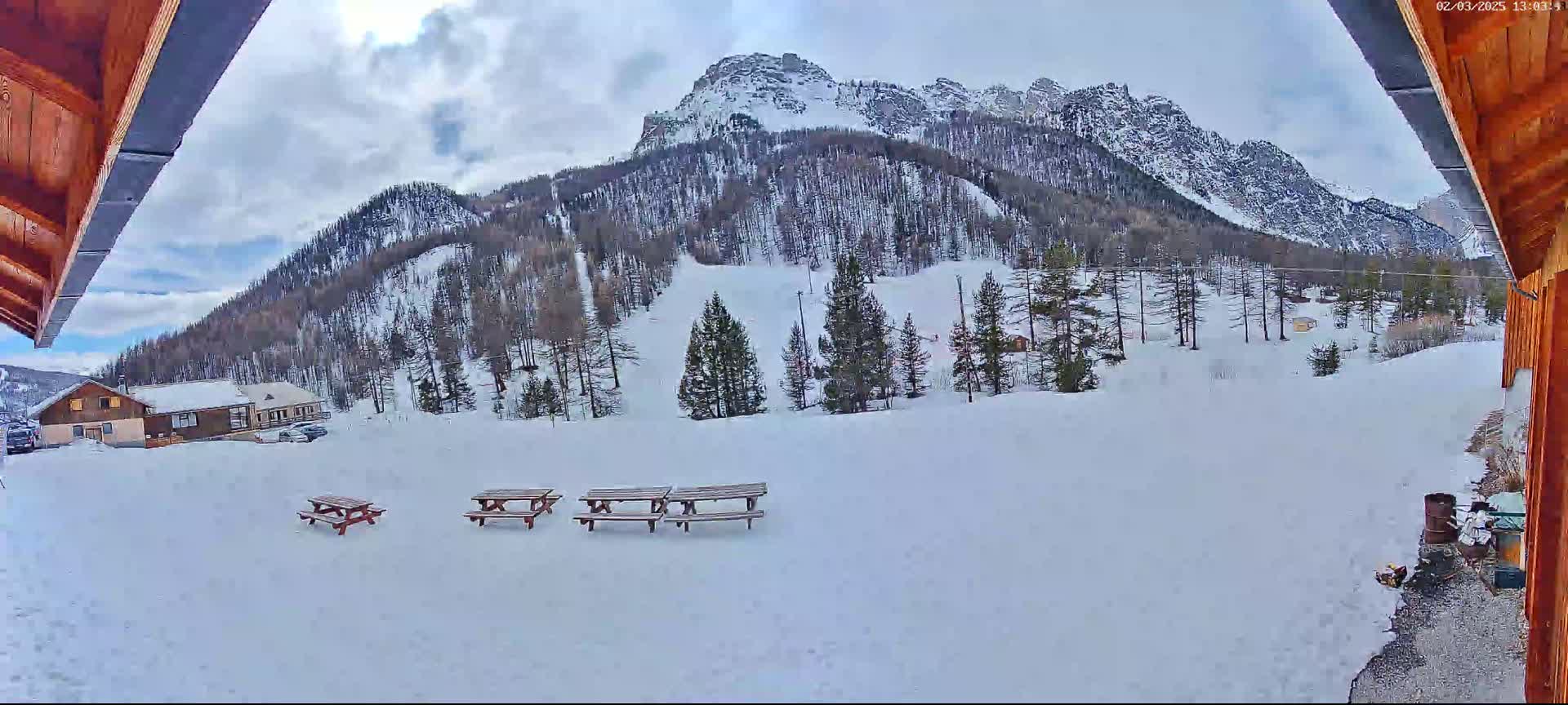 A snowy mountain landscape is viewed from a wooden structure, showing several picnic tables in the foreground and a small building and cars in the midground, under a partly cloudy sky.