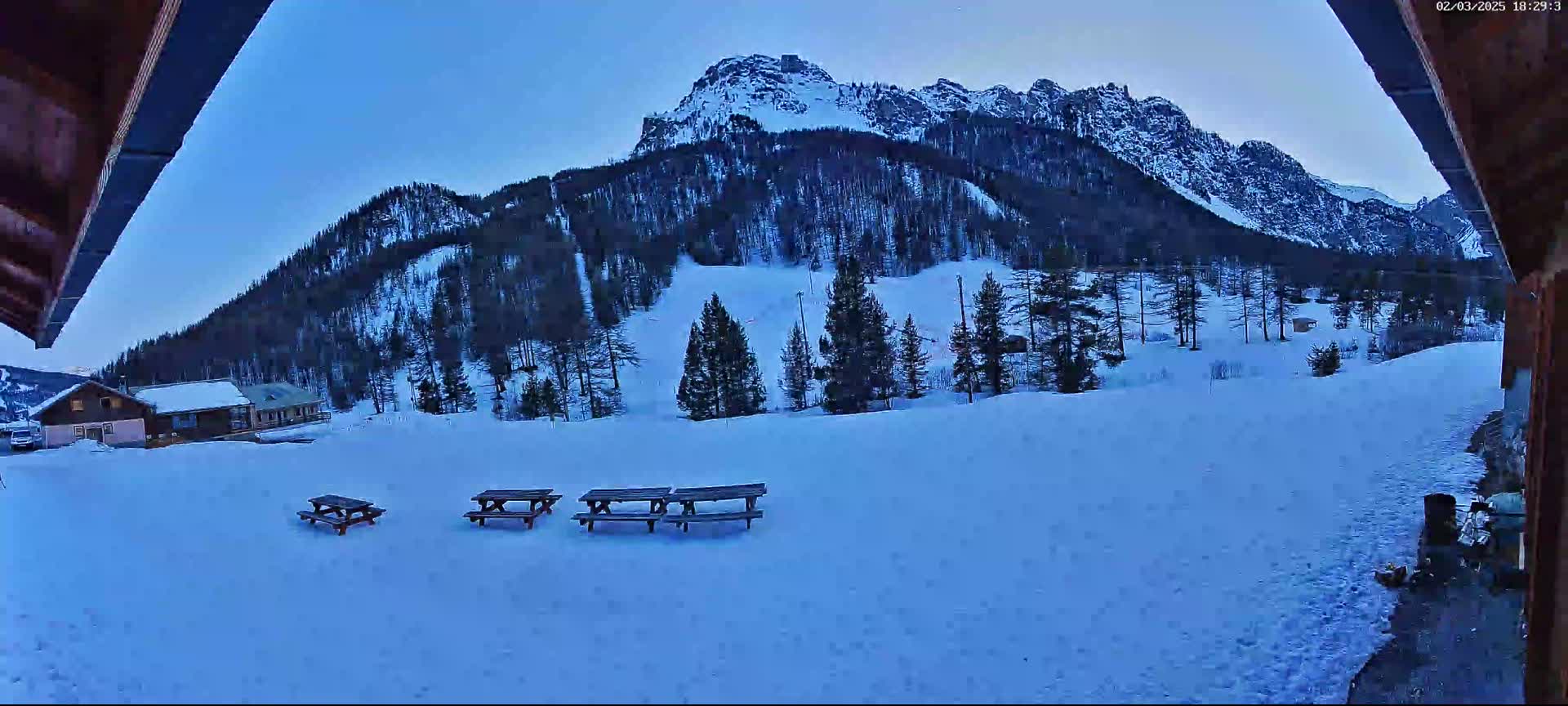 Three picnic tables sit in a snow-covered area overlooking a snow-capped mountain range under a twilight sky.