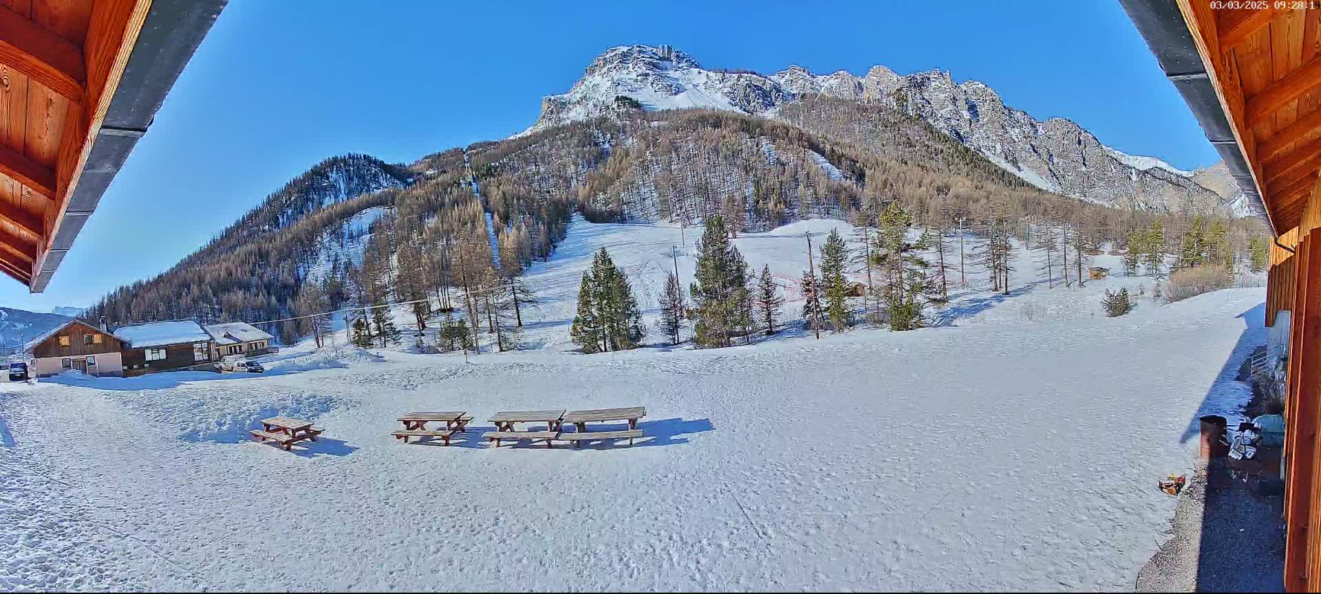 A snow-covered mountainside with several picnic tables in the foreground, viewed from under a wooden roof overhang on a sunny day.