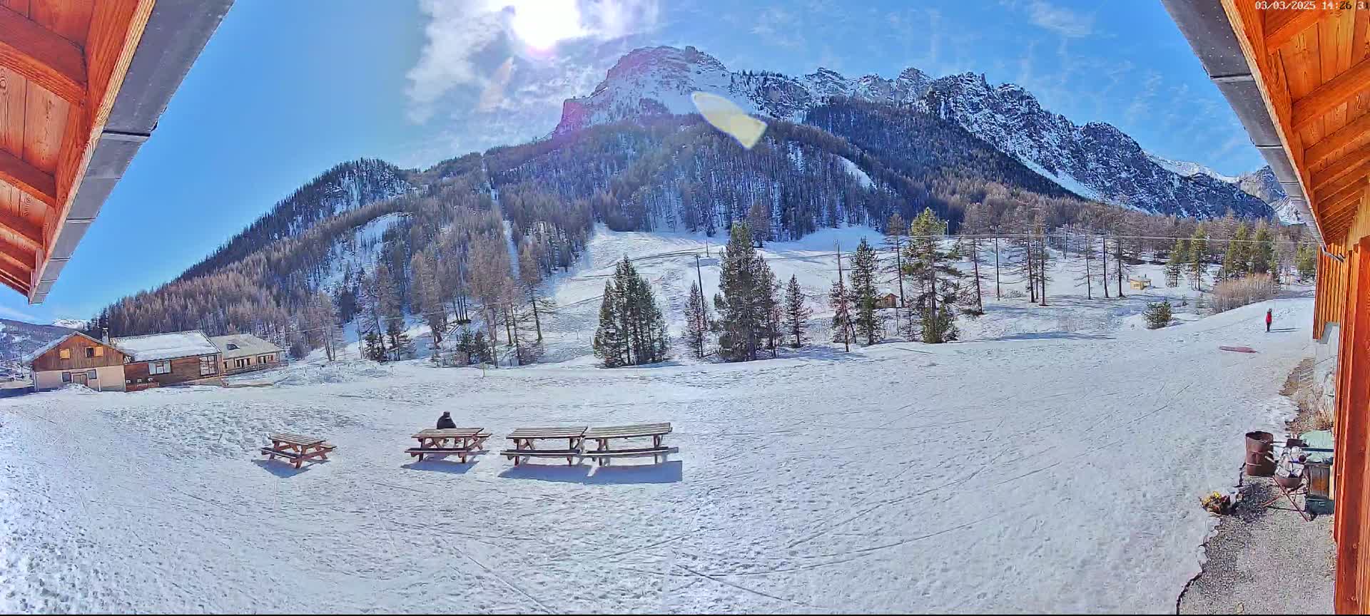 A sunny, snow-covered mountain landscape is seen from a wooden structure, with picnic tables and a person sitting at one in the foreground.