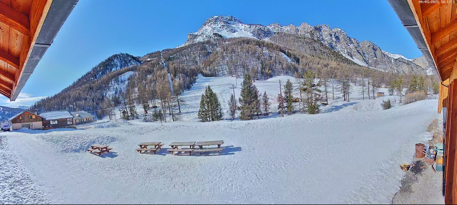 A snow-covered mountainside with several picnic tables in the foreground under a clear blue sky.