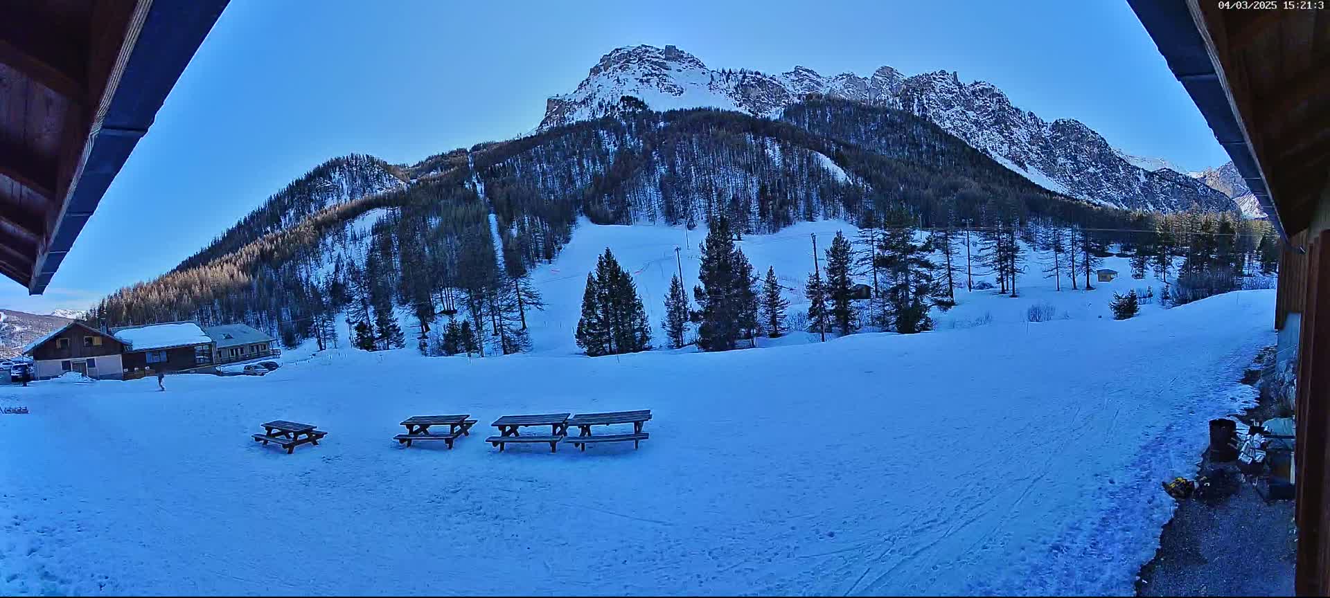 A snow-covered landscape with several picnic tables sits in front of a snow-dusted mountain range under a clear blue sky.