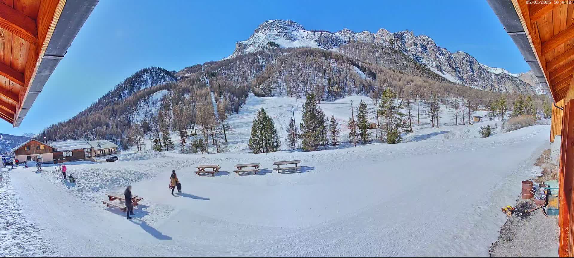 A sunny, snow-covered mountainside with several picnic tables, a few people, and a small building is viewed from a partially enclosed wooden structure.
