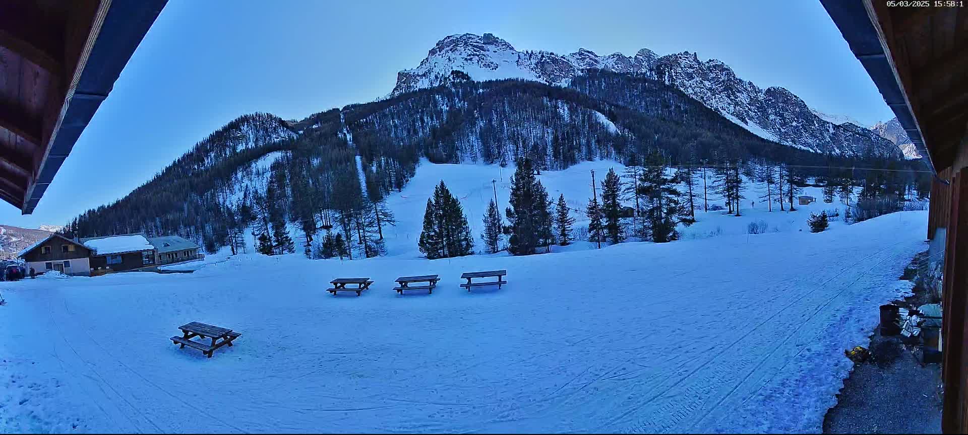 A snow-covered landscape with several picnic tables sits in the foreground, overlooking a mountain covered in dark evergreen trees under a clear blue sky.