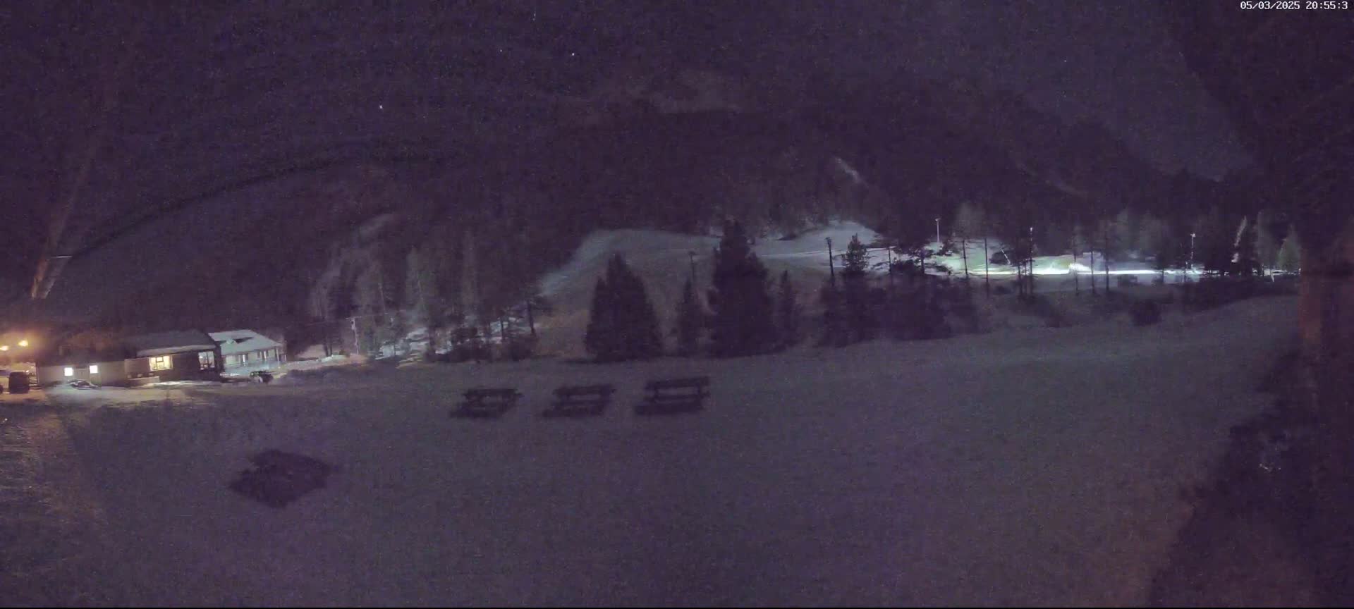 A nighttime, snow-covered scene shows a small building, a few picnic tables, and trees, all set against a dark, mountainous backdrop under a partly cloudy sky.