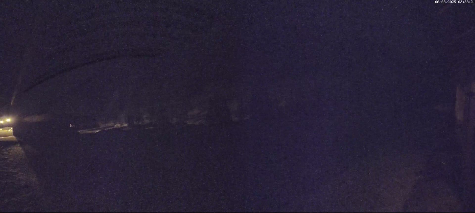 A low-light nighttime image shows a dark, indistinct area that appears to be a roadway or path, possibly lined with trees or shrubs, under a dark sky.