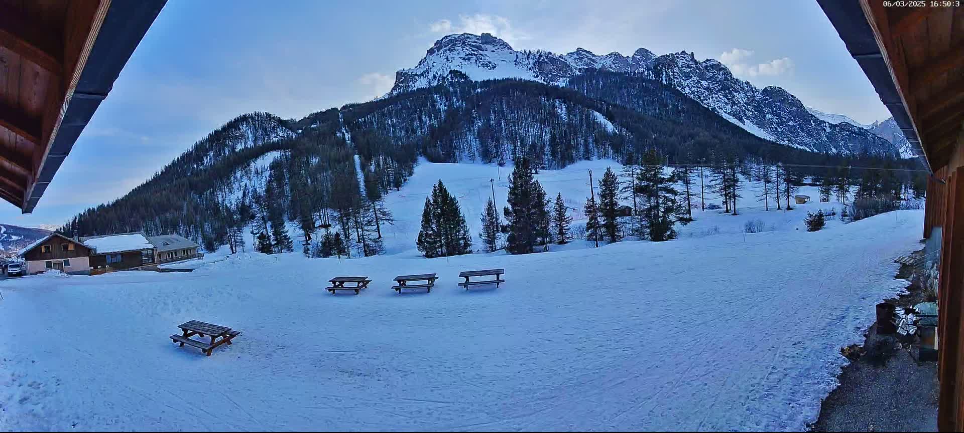 A snow-covered landscape with several picnic tables in the foreground, overlooking a mountain range under a mostly clear sky.