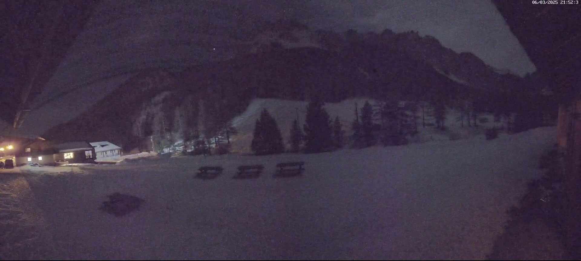 A nighttime, snow-covered scene shows a building, some trees, and picnic tables in front of a mountain under a dark sky.