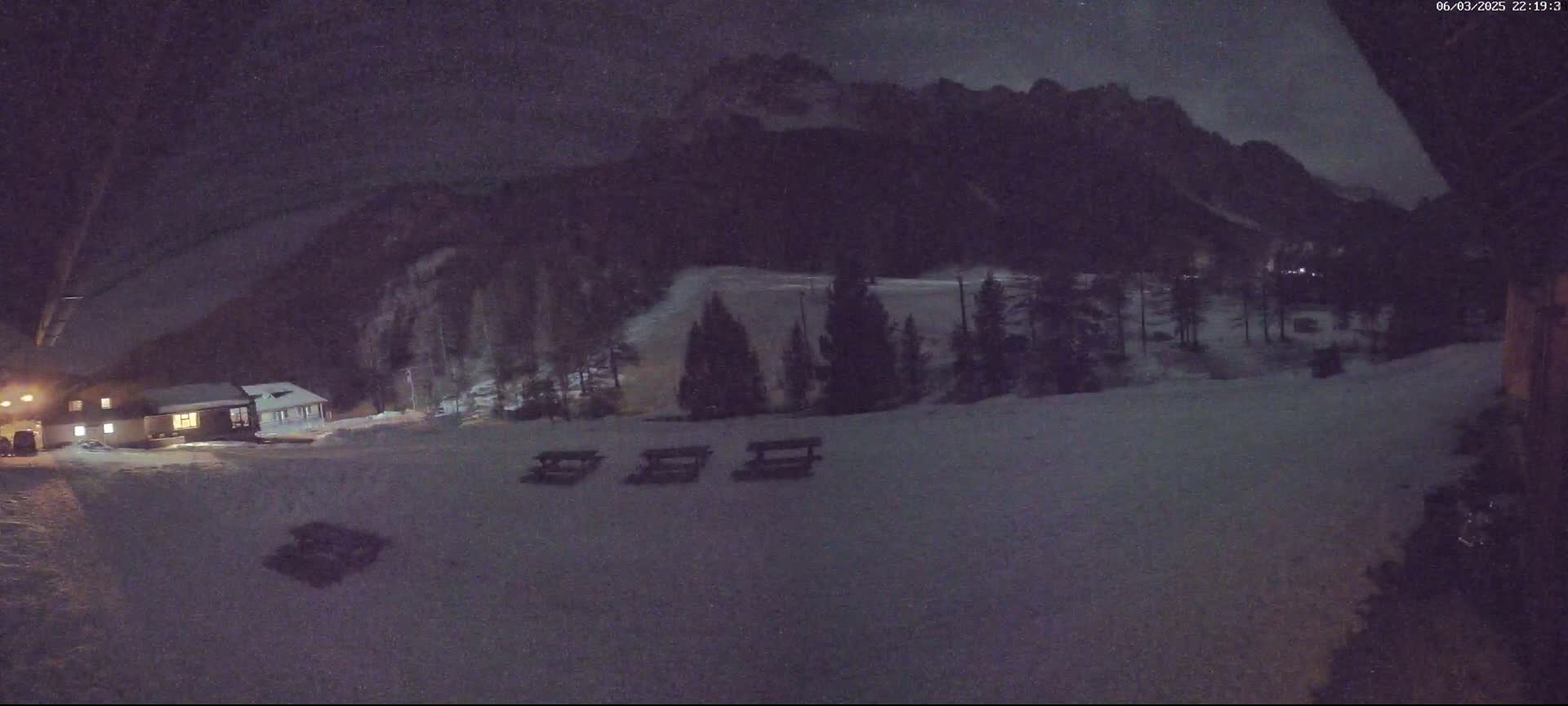 A nighttime, snow-covered landscape shows a building, some picnic tables, and a dark mountain range in the background under a lightly clouded sky.
