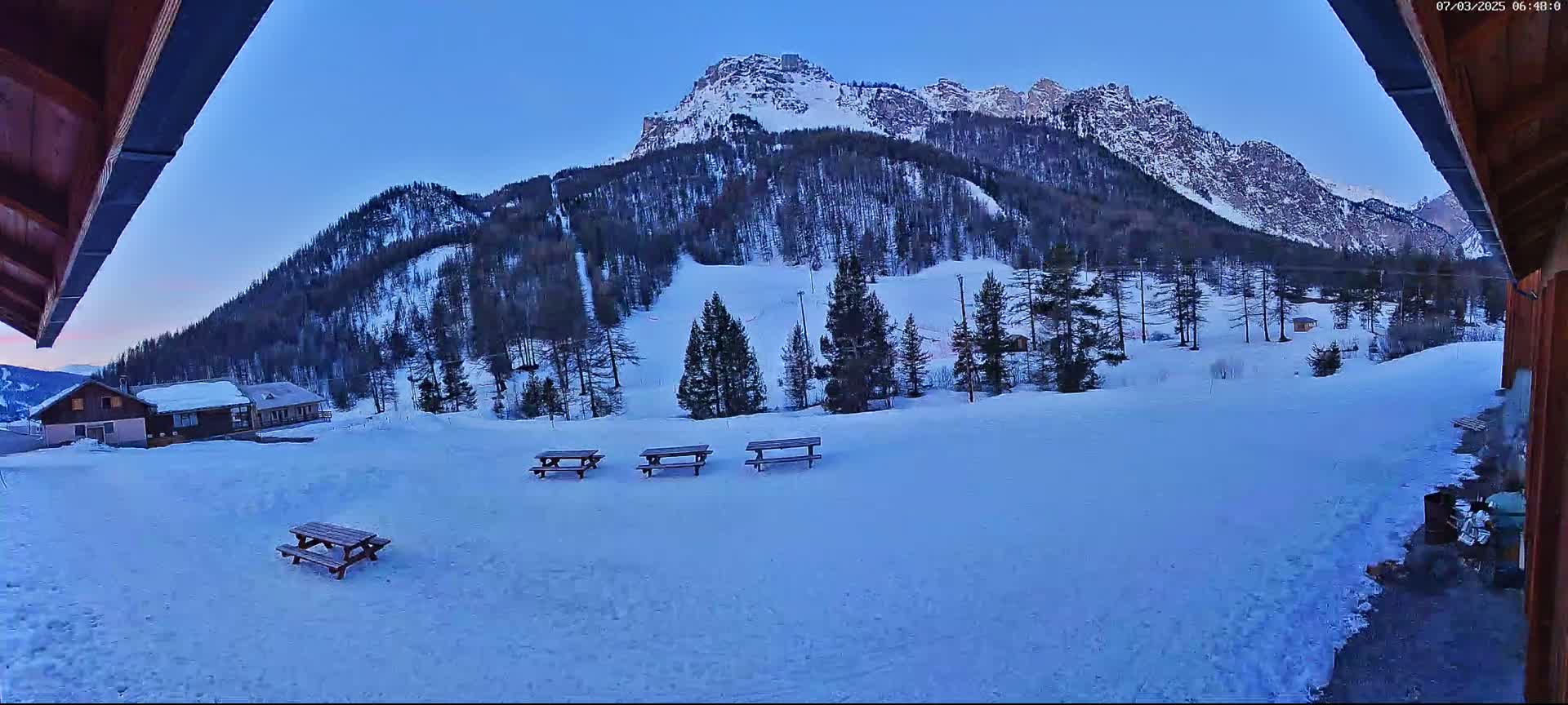 A snow-covered landscape with several picnic tables in the foreground, overlooking a mountain range under a clear, twilight sky.