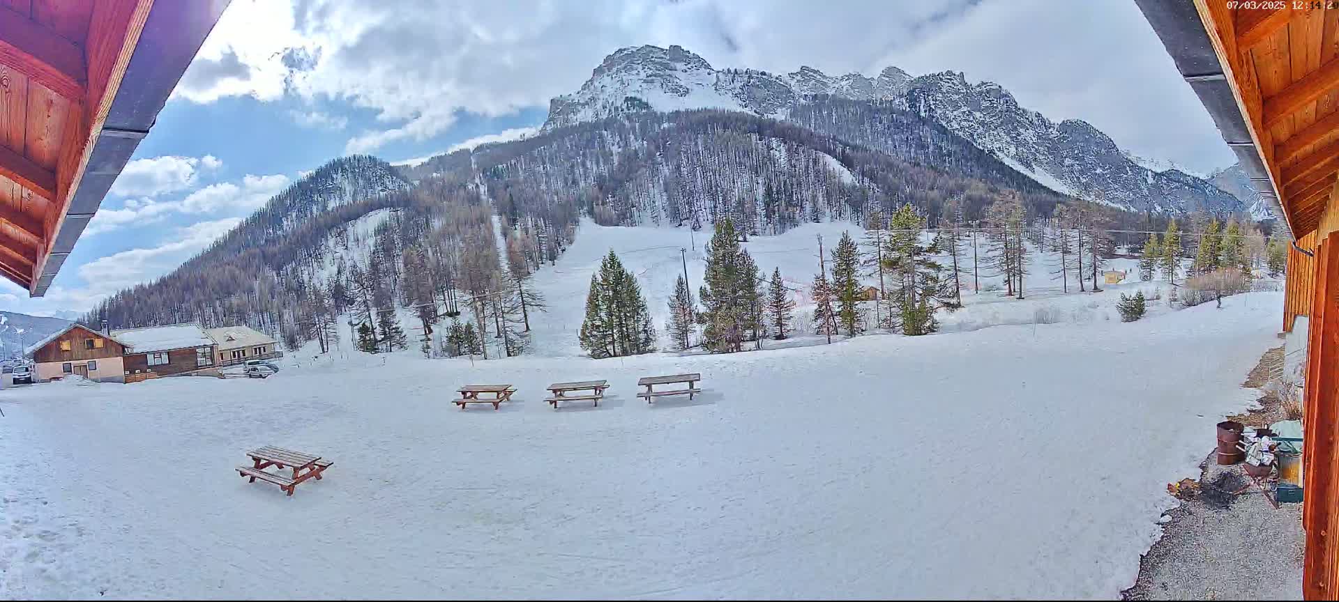 A snow-covered landscape with several picnic tables, a few buildings, and a mountain range in the background under a partly cloudy sky.