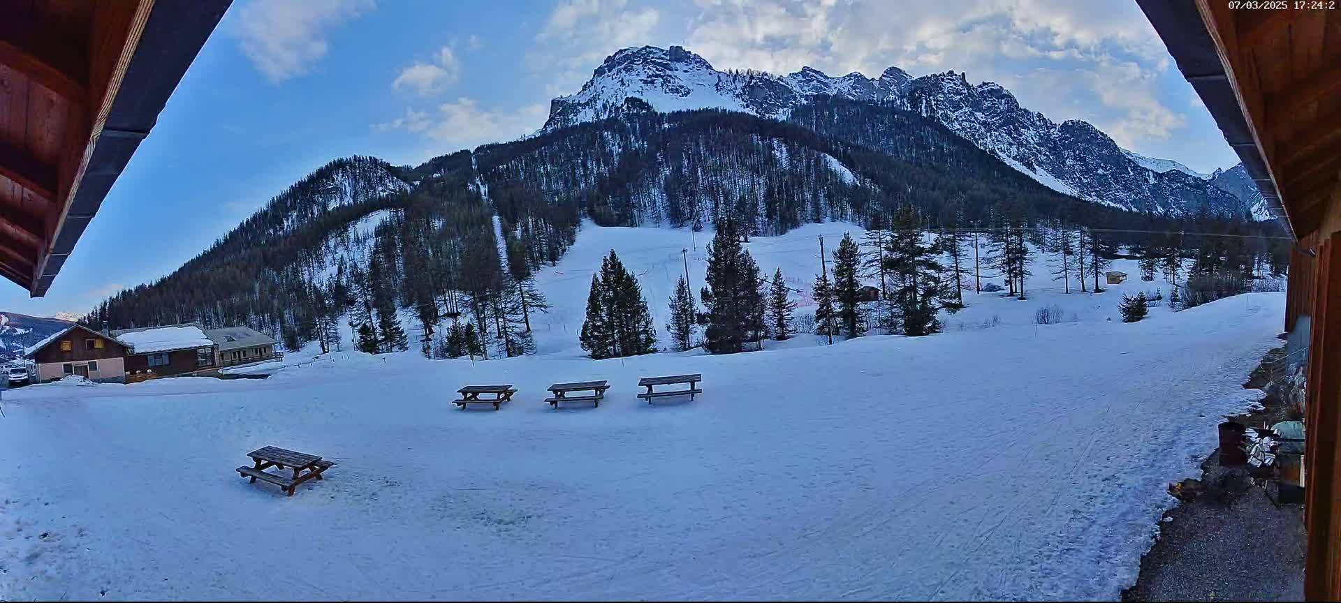 A snow-covered landscape with several picnic tables, a few buildings, and a mountain range in the background under a partly cloudy sky.