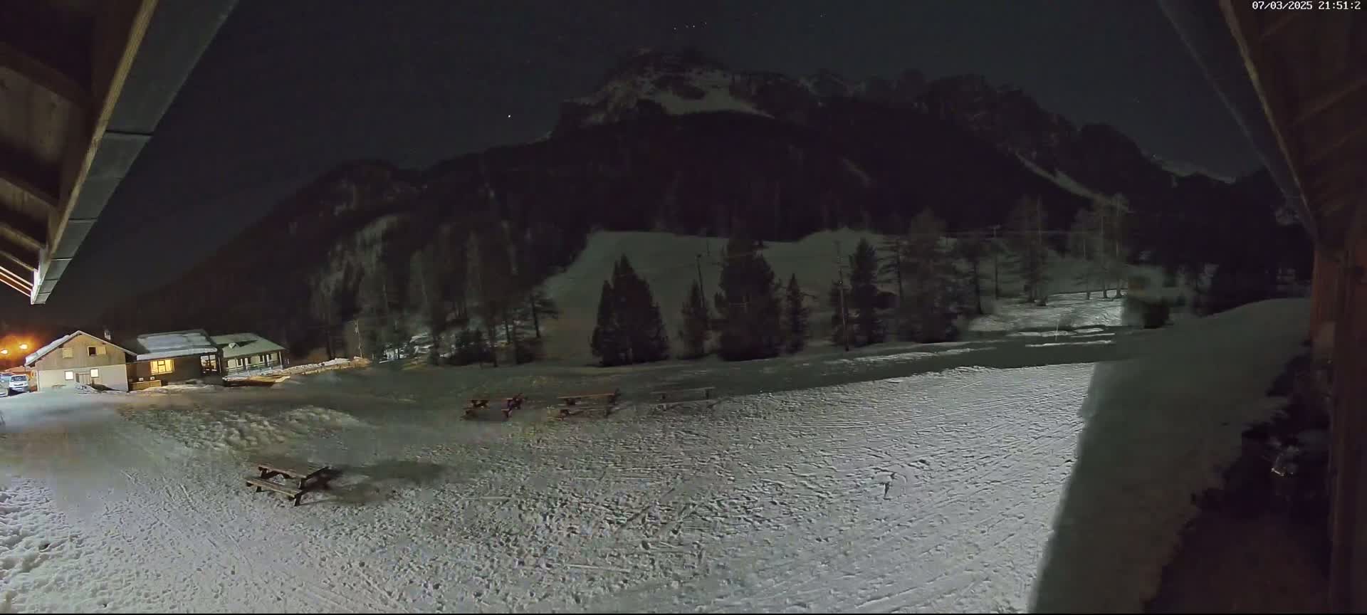 A snow-covered landscape at night, illuminated by a faint light, shows a building, some picnic tables, and dark trees against a backdrop of mountains under a mostly clear, dark sky.