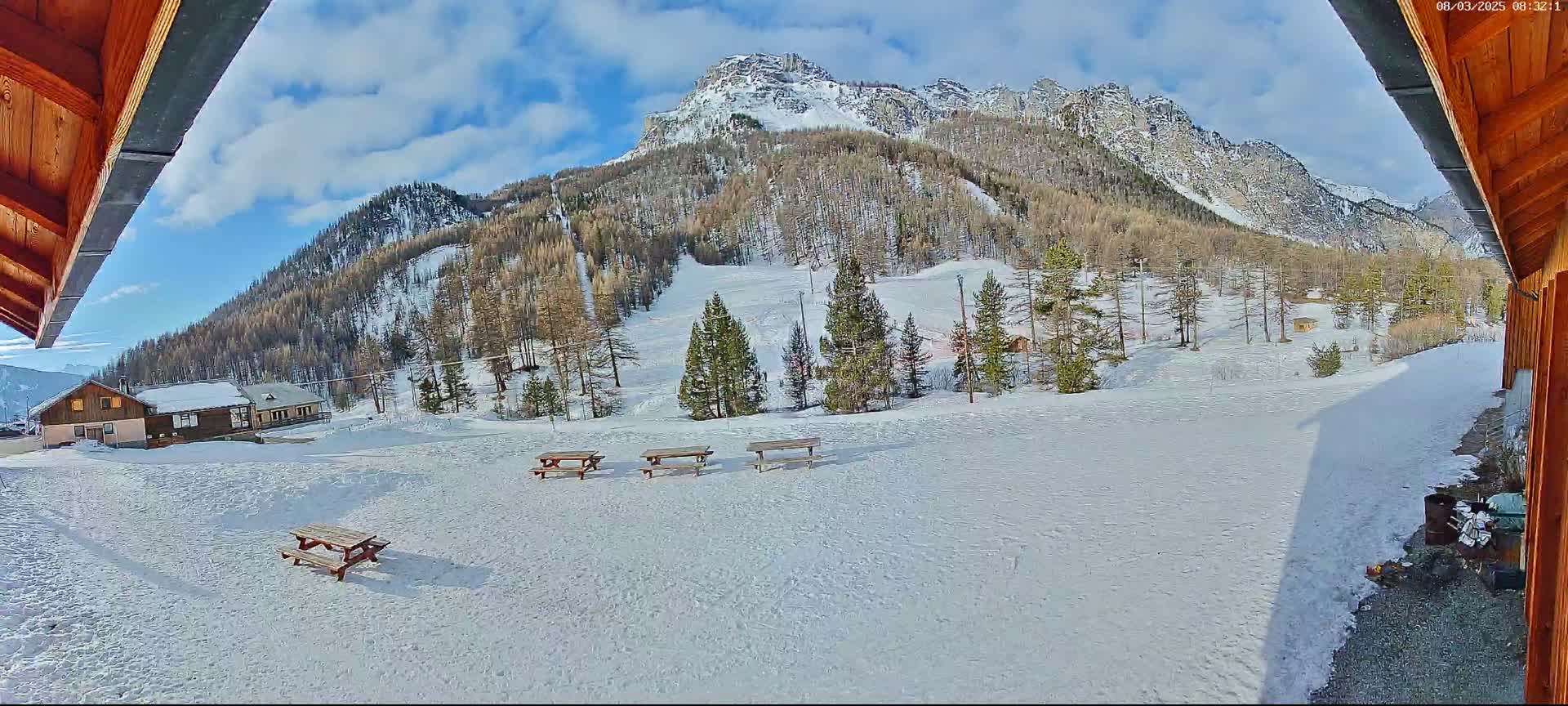A snowy, mountainous landscape is viewed from a wooden structure, featuring several picnic tables in the foreground and a small building in the mid-ground.